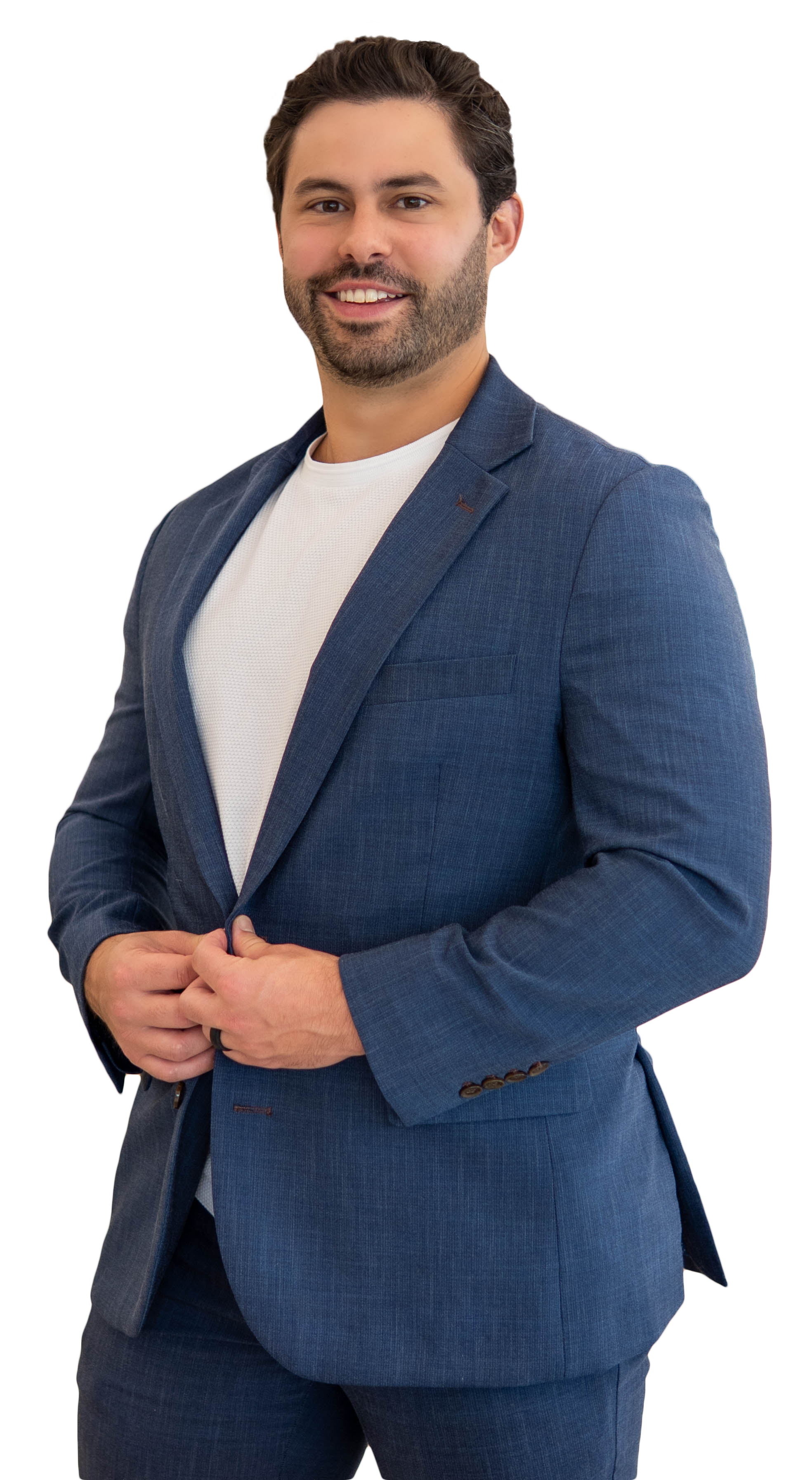 A man with dark hair and a beard dressed in a blue suit with a white shirt underneath, smiling and standing with his hands together in front of him.
