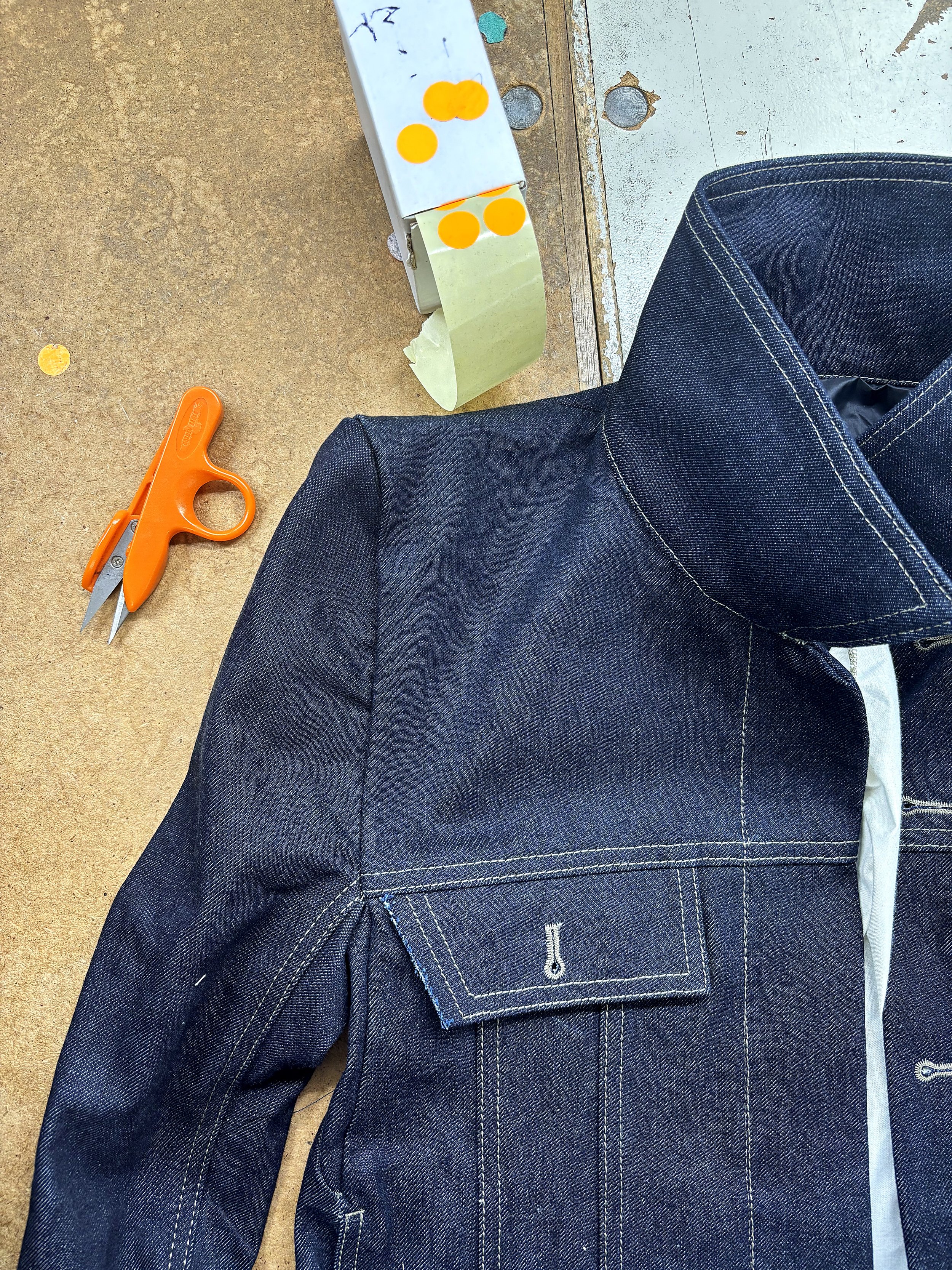 Giacca da lavoro in denim scuro con cuciture bianche, forbici arancioni e nastro adesivo su un banco da lavoro di Lineare51, servizi per l'abbigliamento e confezione Rovigo.