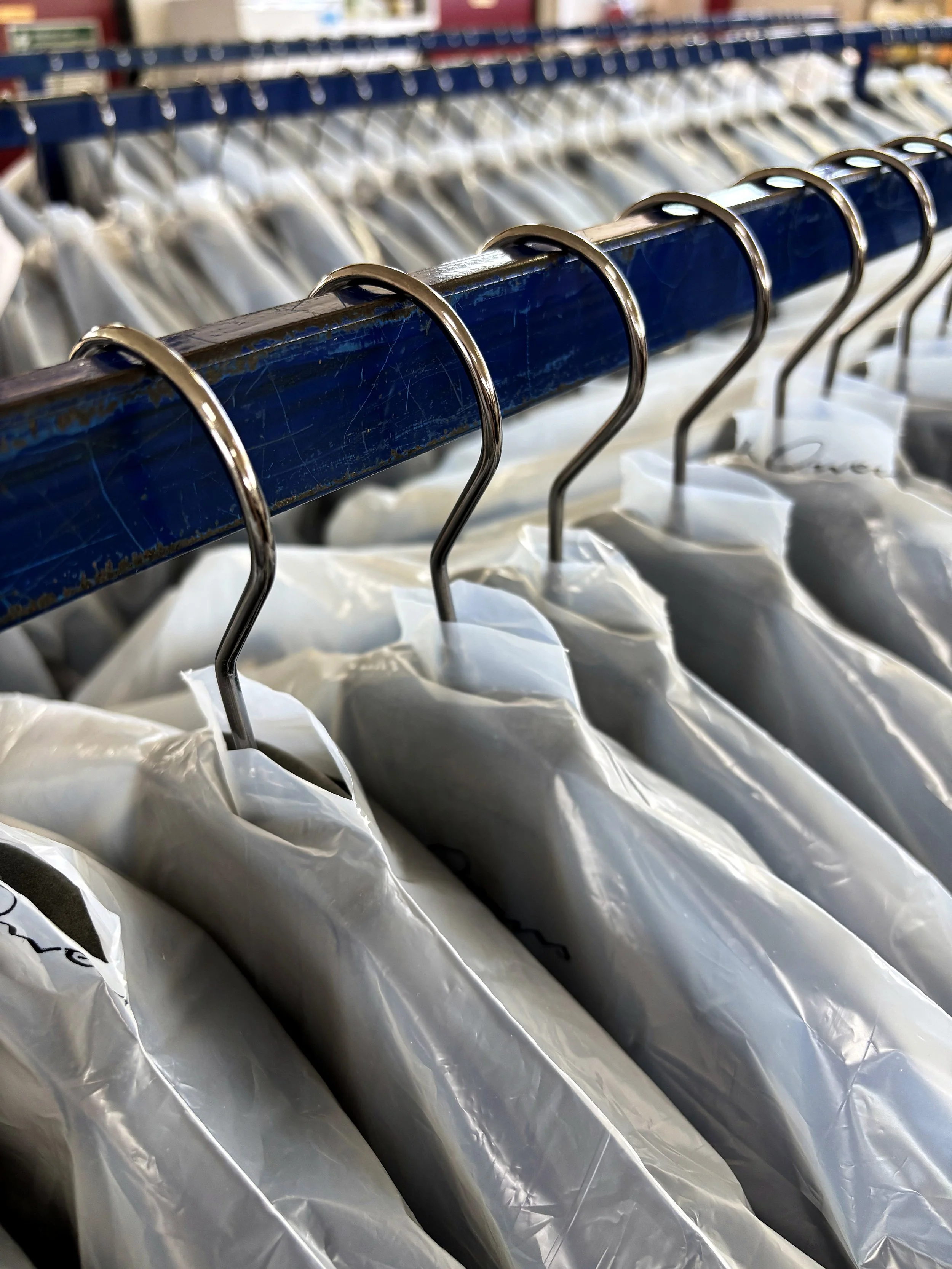Primo piano di grucce metalliche blu appese a un’asta, con indumenti bianchi protetti da sacchetti di plastica di Lineare51, servizi per l'abbigliamento e confezione Rovigo.