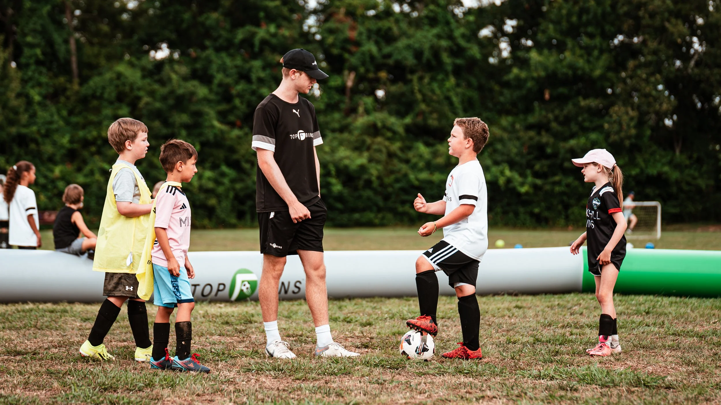 Children and an adult coach on a soccer field, three children are standing, one is kneeling, and the coach is talking to a girl who is holding a soccer ball. Other children are in the background with a goal post and trees.