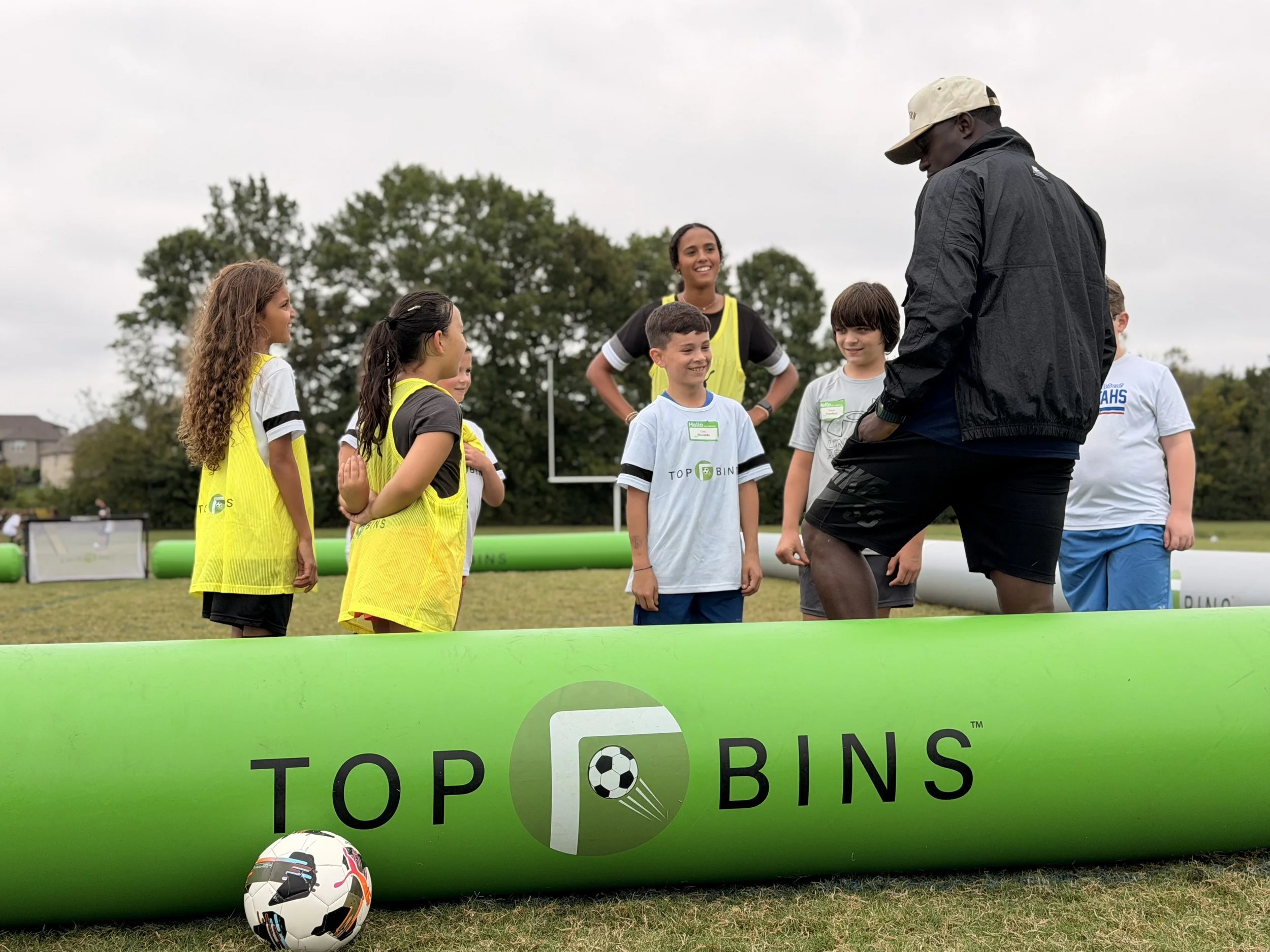 A group of children and a coach standing on a soccer field during the day, engaging in an activity related to trash disposal and environmental awareness, with a prominent green banner displaying a message about stopping trash and a soccer ball on the grass.