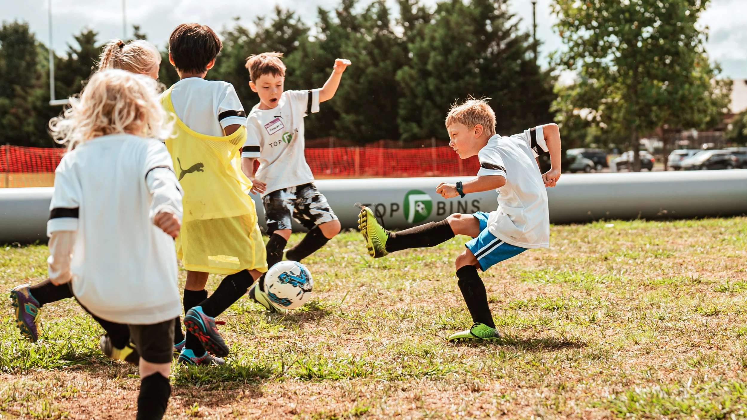 Children playing soccer outdoors on a grassy field with trees and a red barrier in the background.