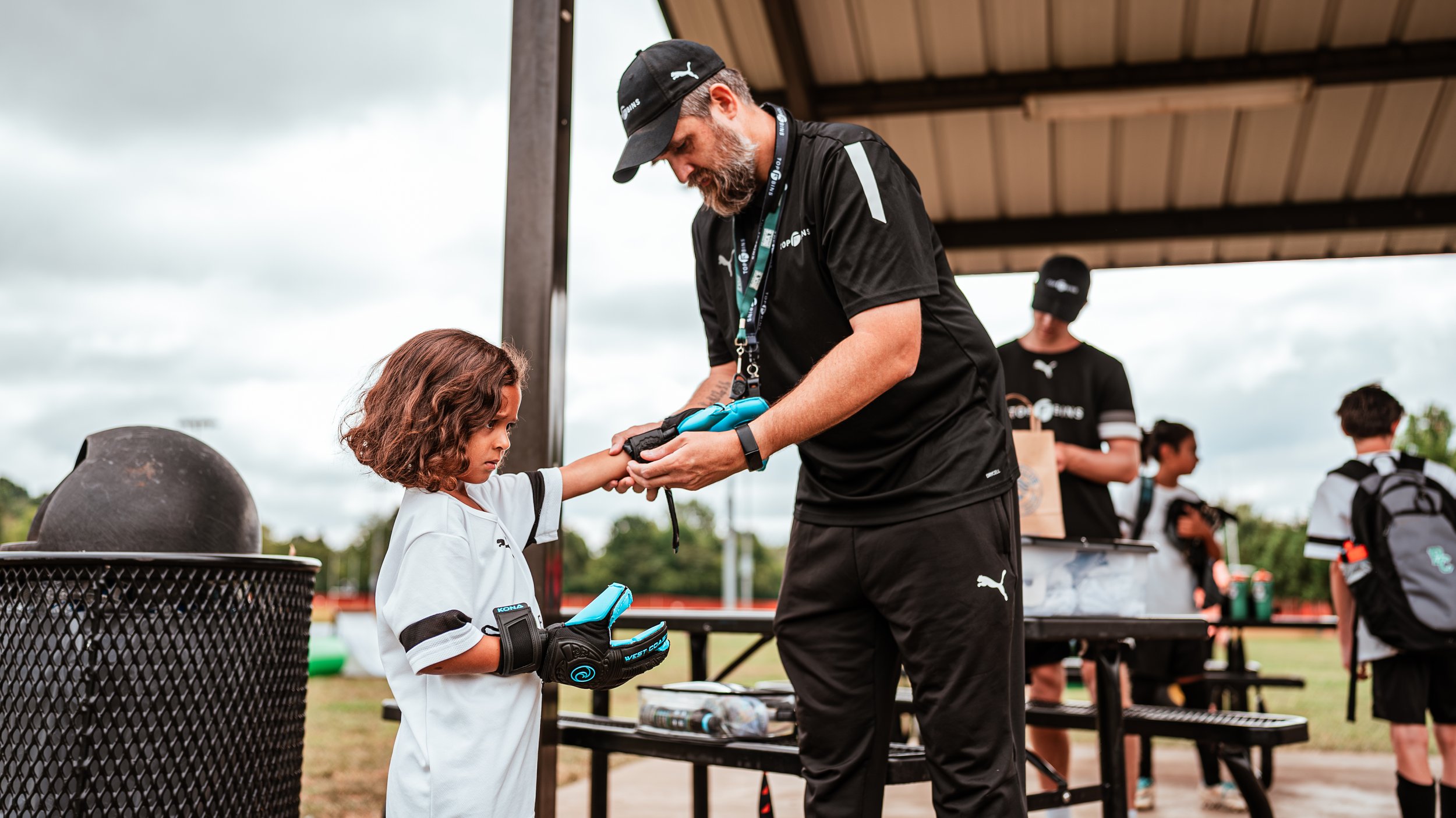 A young girl in a hockey uniform receives a gift from a man in a black Puma shirt and hat, under a pavilion with other children and adults in the background.