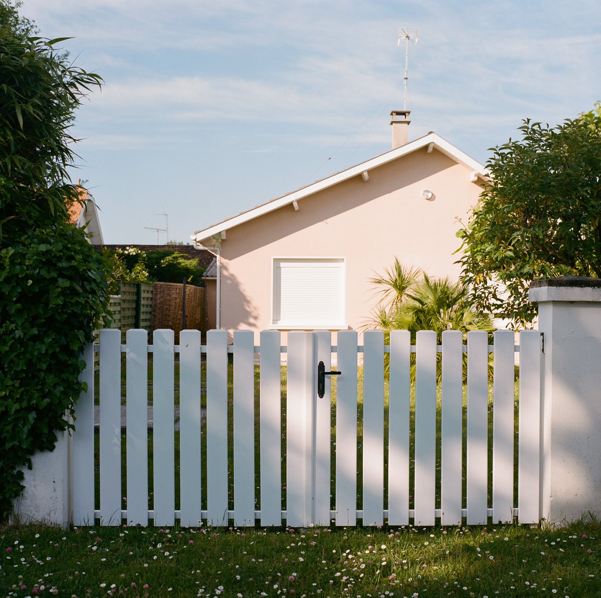 Une maison beige vue de l'extérieur, entourée d'une clôture blanche, avec des arbres verts dans le jardin et un ciel bleu en arrière-plan.