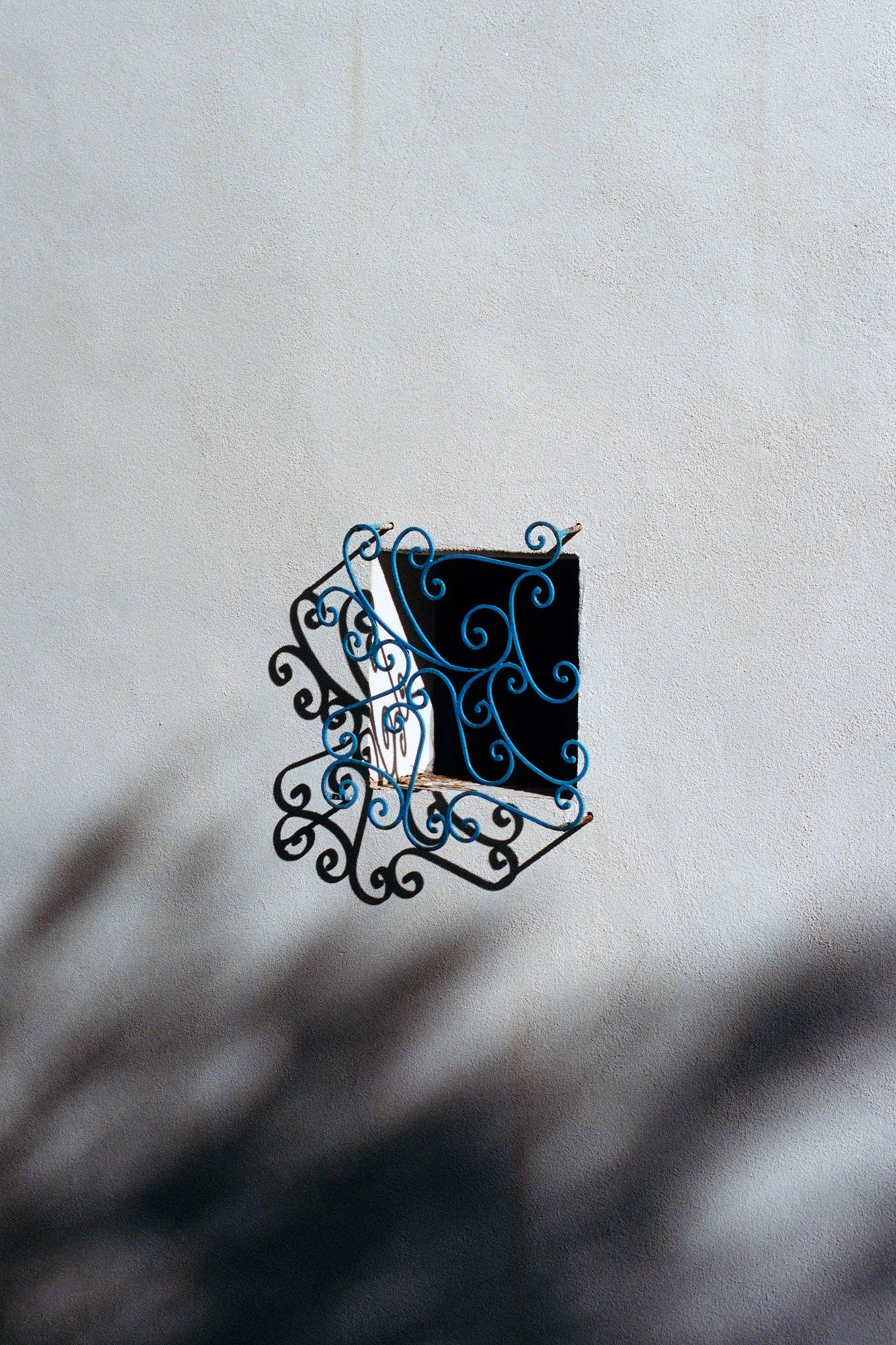 Une chaise en fer forgé avec un motif de spirale, fixée à un mur blanc, projette une ombre sur le mur.