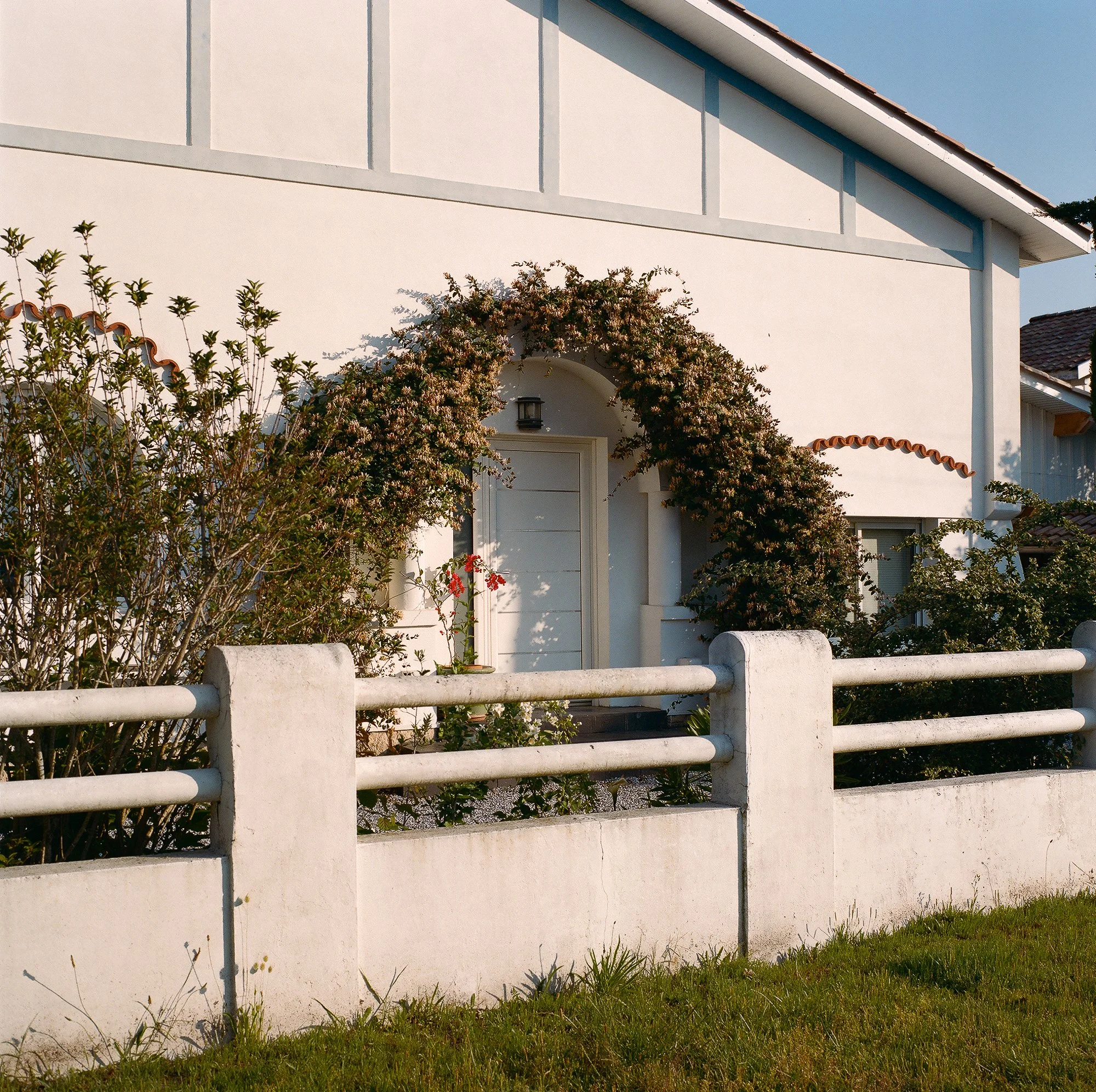 Une maison moderne blanche avec un toit en tuiles rouges, entourée d'un mur en béton blanc et de végétation, sous un ciel clair.