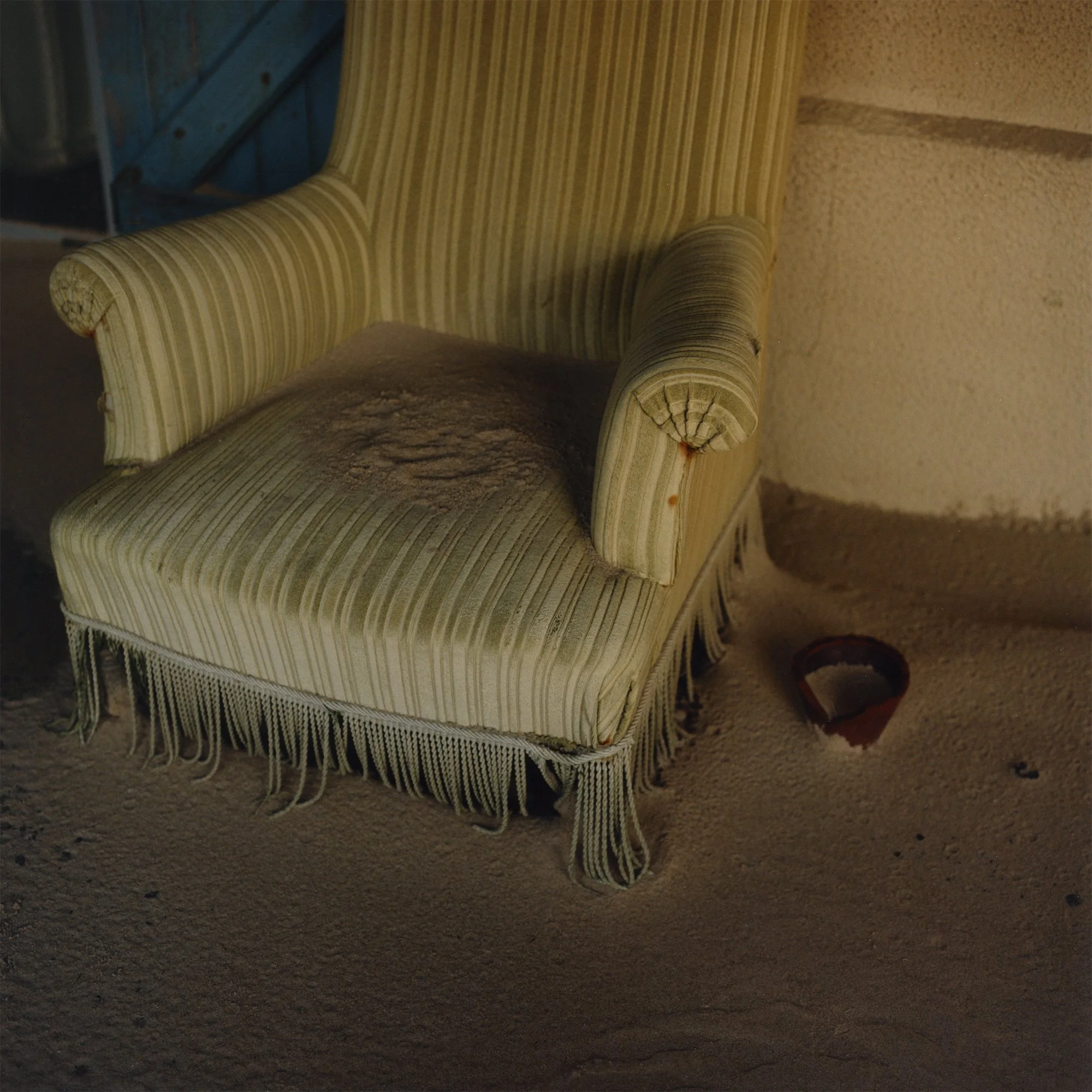 Une vieille chaise rembourrée en tissu à rayures jaunes et blanches avec une frange en bas, située sur un sol en béton, avec un pot de fleurs en terre cuite à droite