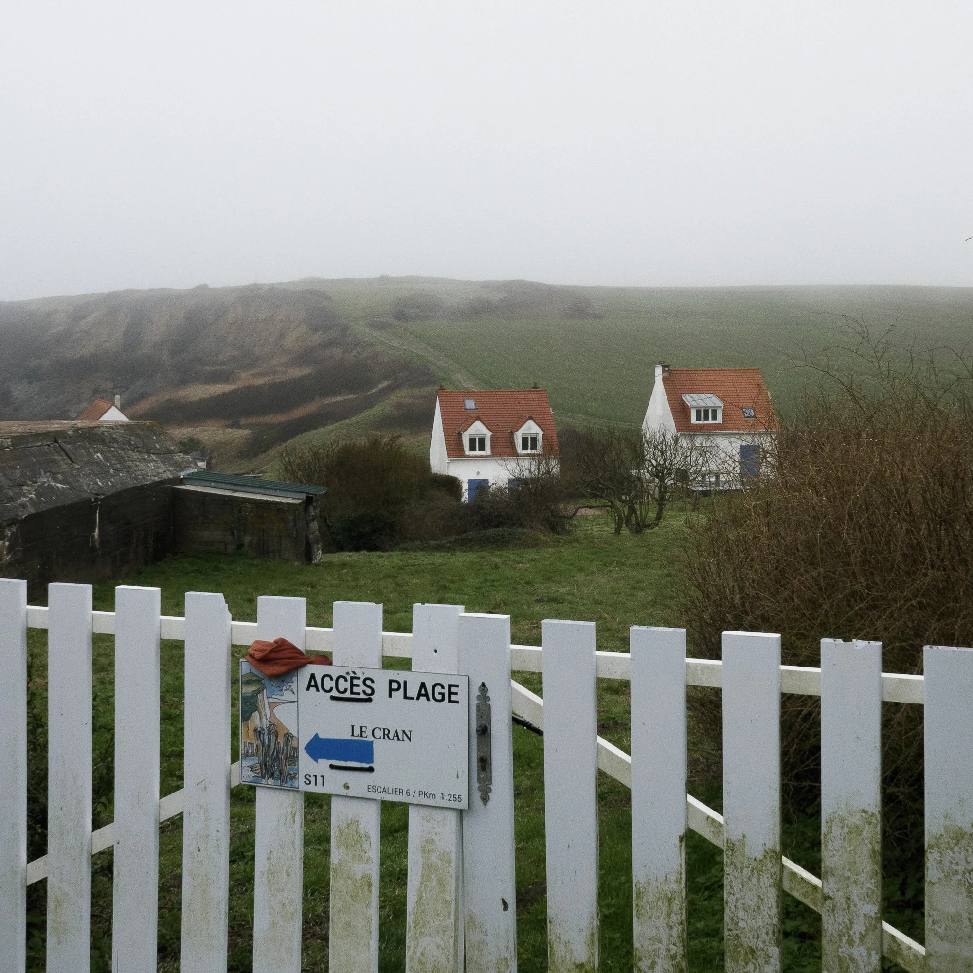 Une barrière blanche avec un panneau d'avertissement indiquant l'accès à la plage. En arrière-plan, il y a des maisons avec des toits en tuiles rouges et un paysage de collines verdoyantes sous un ciel nuageux.