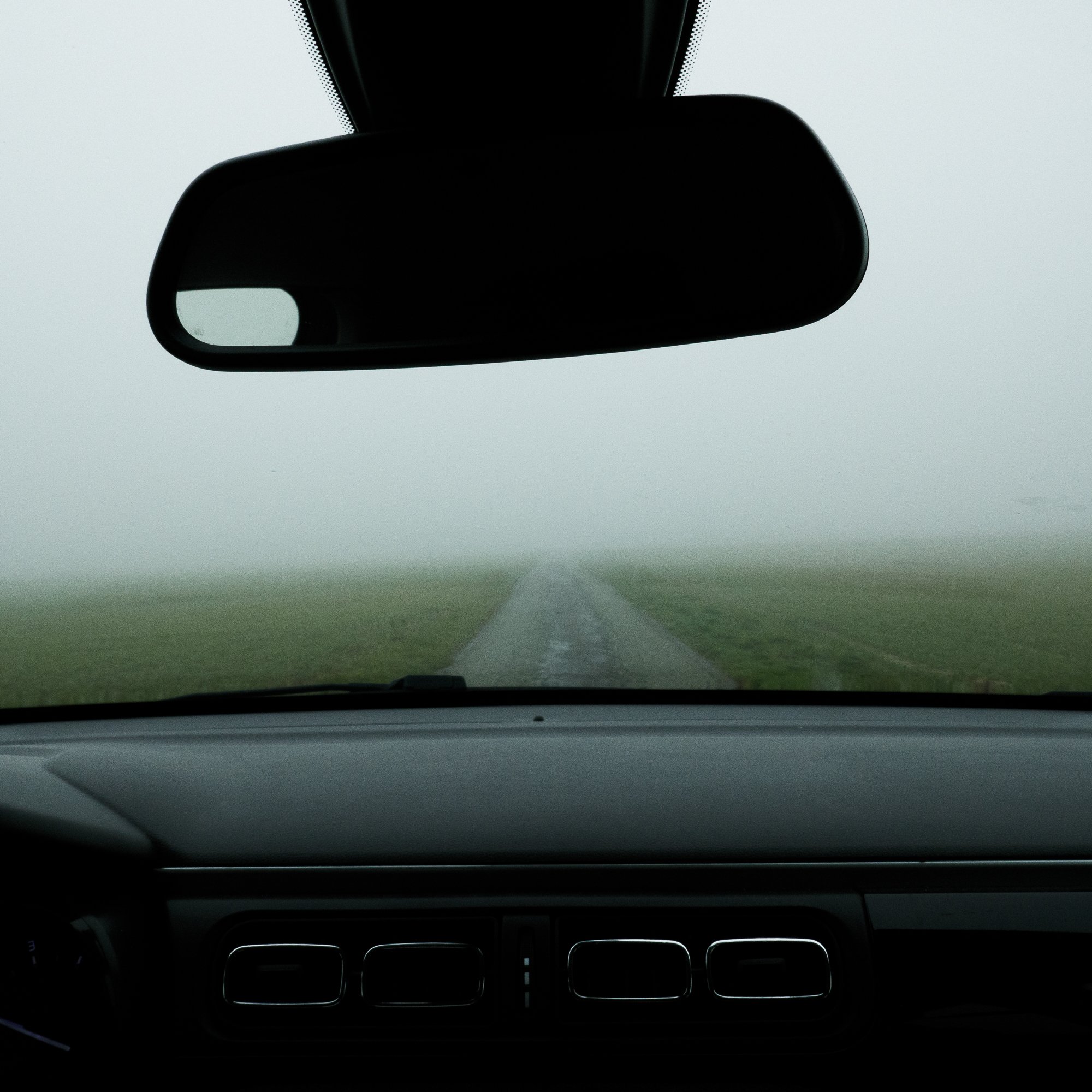 Vue de l'intérieur d'une voiture montrant le rétroviseur, le tableau de bord et la vue à travers le pare-brise vers une route verte dans le brouillard.