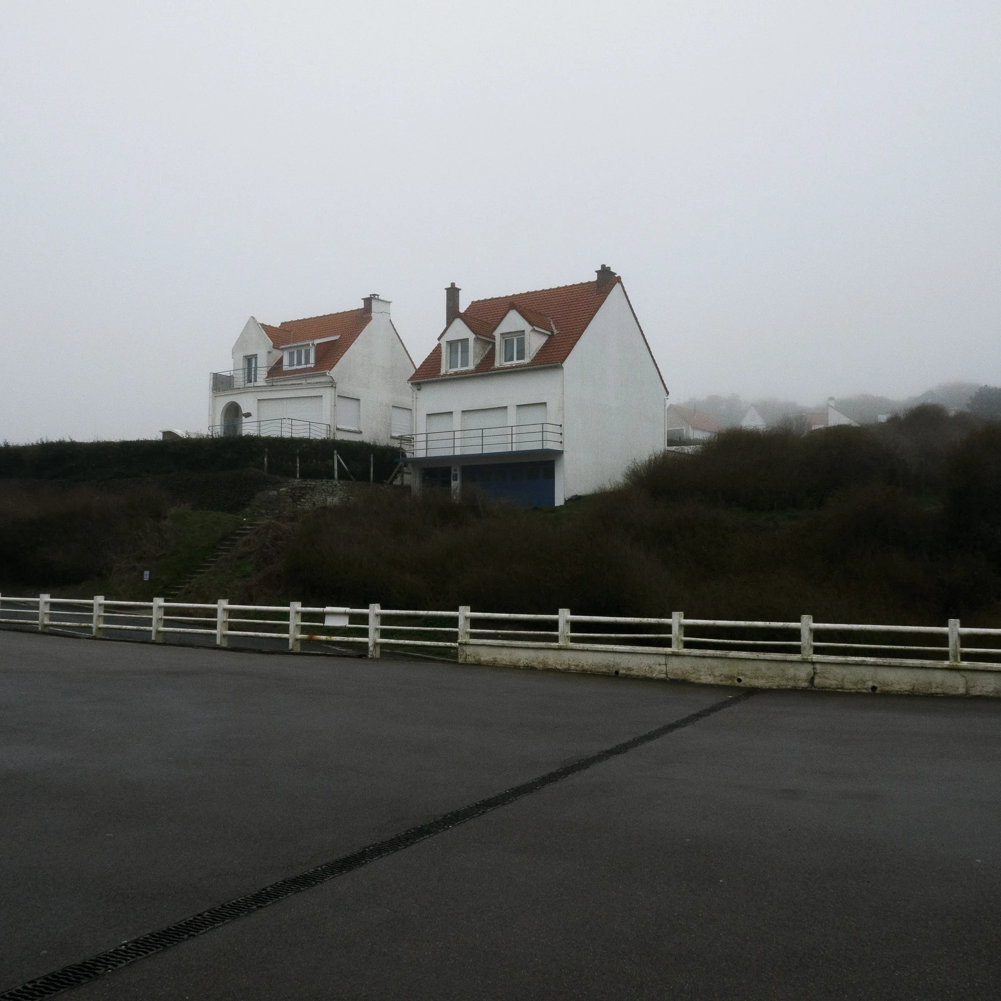 Vue de maisons blanches avec toits en tuiles rouges sous un ciel brumeux, bordée d'une route avec une barrière blanche en premier plan.
