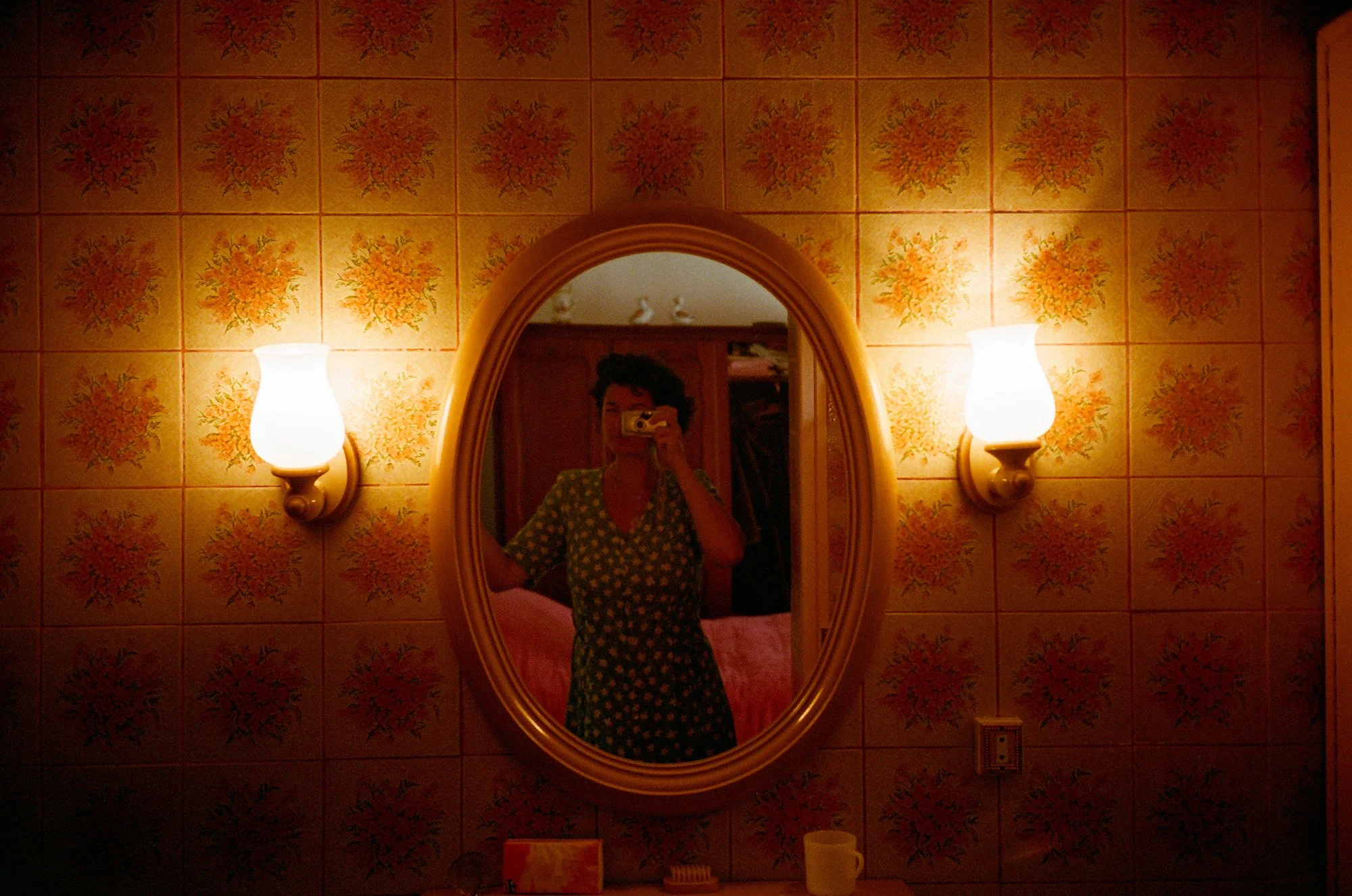 Un miroir oval sur un mur tapissé de carreaux orange avec motif floral, éclairé par deux appliques murales, une personne avec un casque et un vêtement à motifs est en train de prendre une photo d'elle-même dans le miroir.
