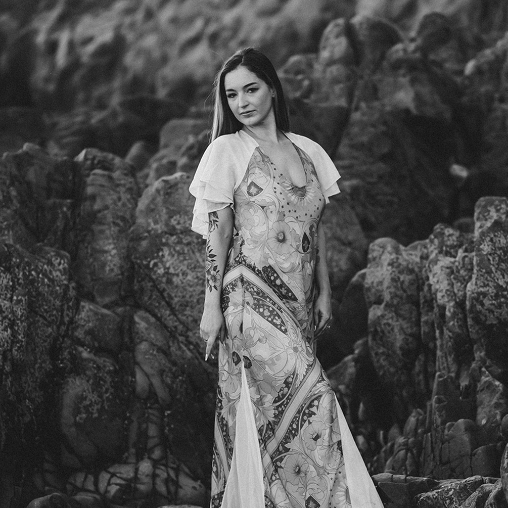 A woman with long dark hair standing on rocks near the ocean, wearing a flowing dress with a floral and geometric pattern.