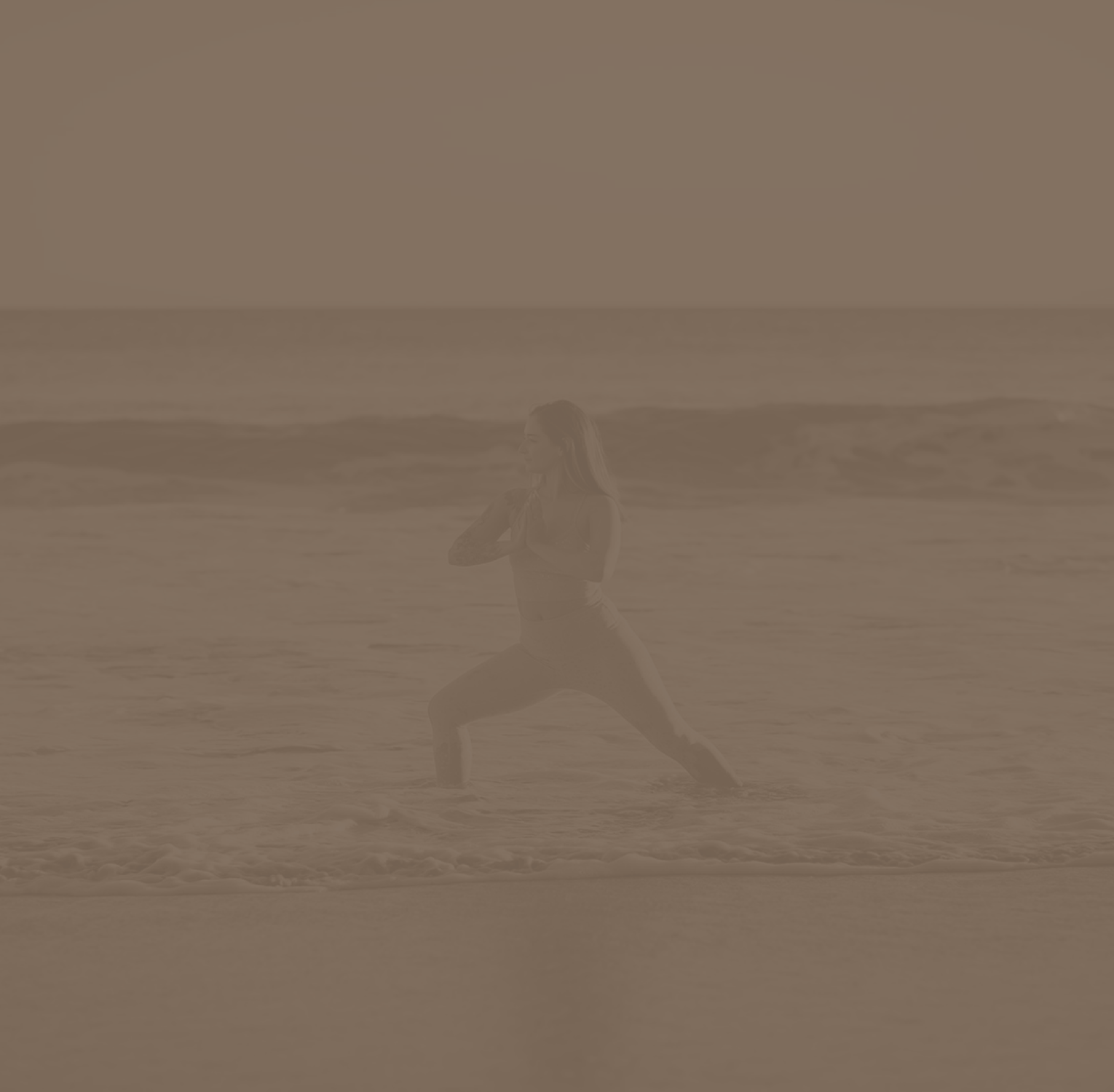 A woman practicing yoga in a lunge pose on a beach with the ocean and a clear sky in the background.