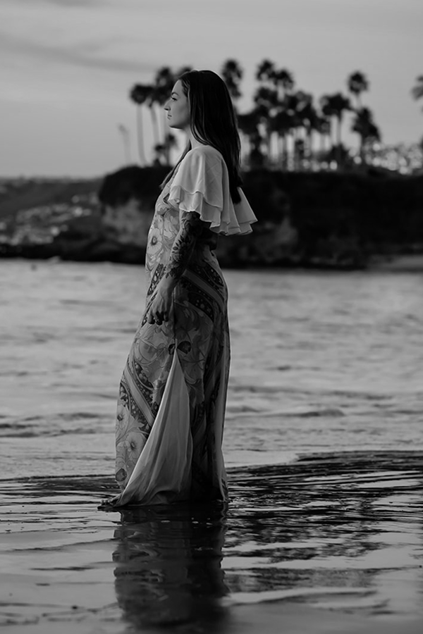 A woman standing in shallow water on a beach, looking sideways, with palm trees and a rocky cliff in the background, black and white photo.