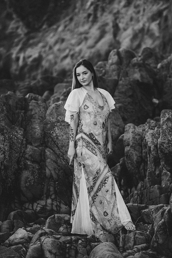 A woman standing on rocks near a rocky shoreline or hillside, wearing a floral dress with flowing fabric and looking to the side.