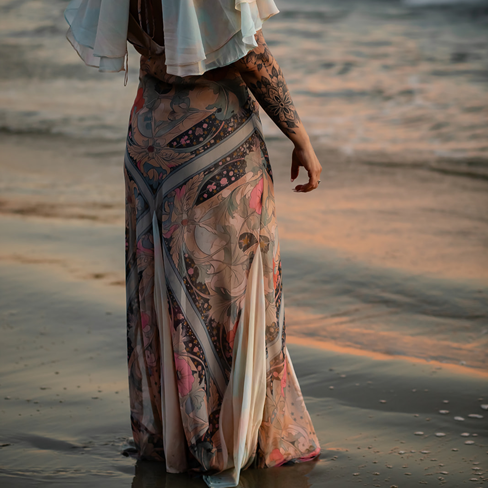 A person walking on the beach at sunset, wearing a flowing, patterned skirt and a light-colored top, with visible tattoos on their arm.