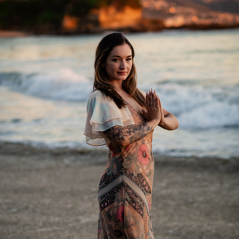 A woman standing on the beach near the water, with waves in the background during sunset, holding her hands together in a prayer pose.
