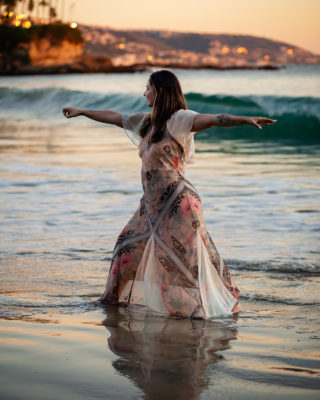 A woman in a flowing floral dress stands in the ocean with arms outstretched during sunset.