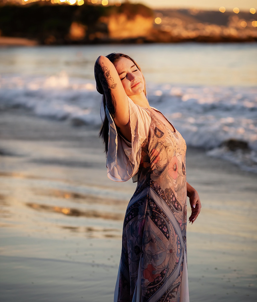 A woman with long dark hair standing on a beach at sunset with her eyes closed and one arm raised, wearing a flowing, patterned dress.
