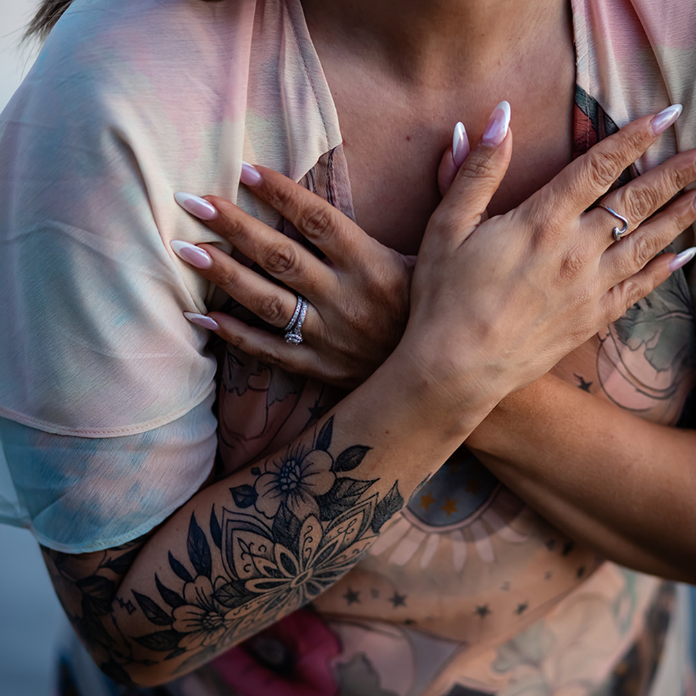 Close-up of a person with tattoos crossing their arms over their chest, wearing rings and long manicured nails.