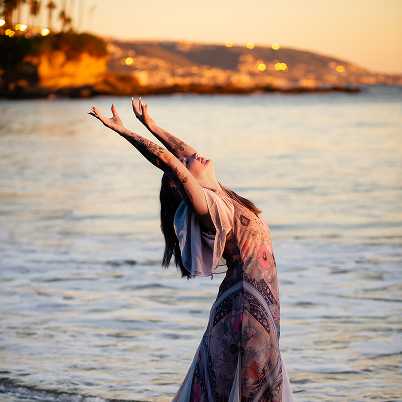 A woman with tattoos on her arms is standing in shallow water at sunset, stretching her arms upwards with her face turned towards the sky, near a coastal landscape with hills and lights in the background.