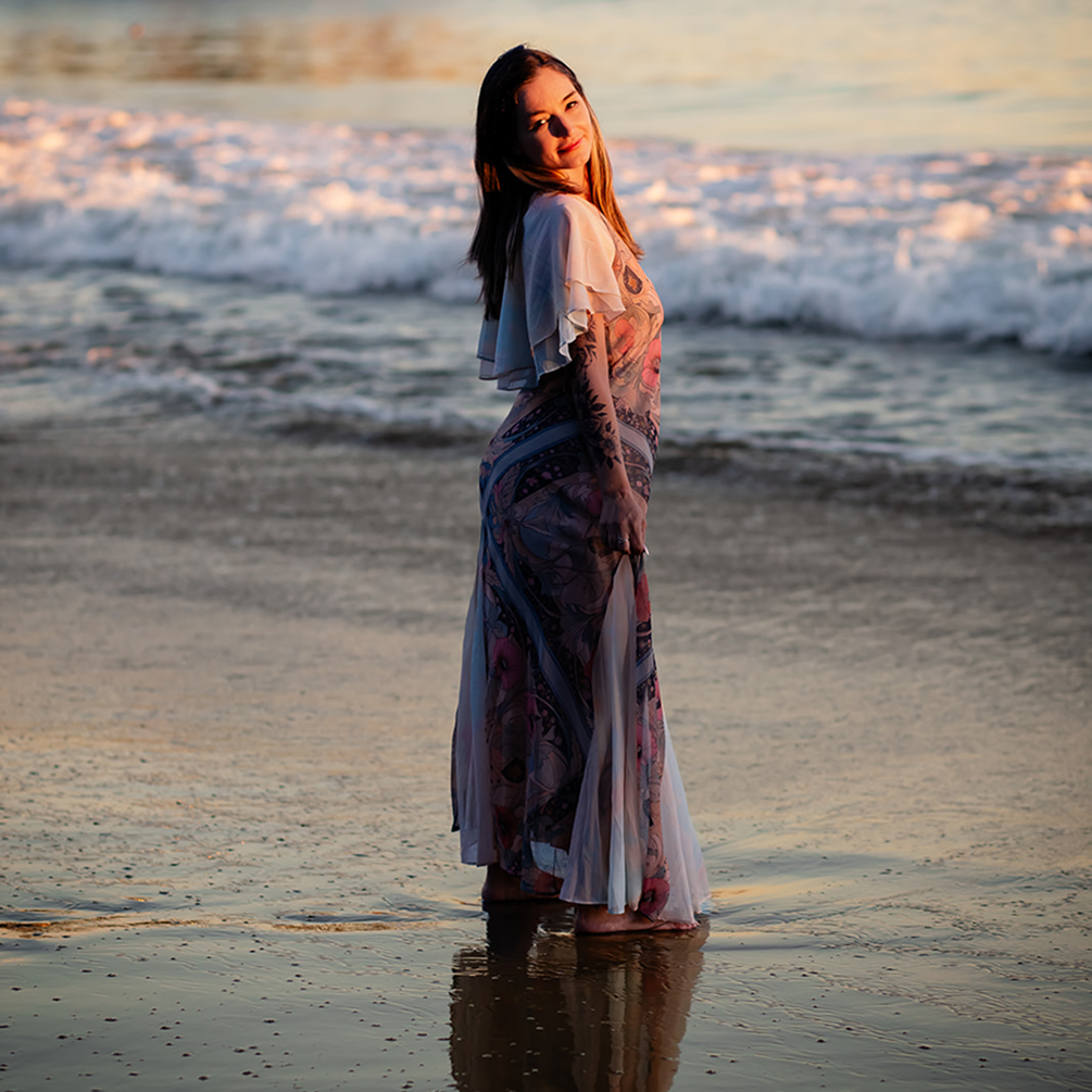A woman in a floral dress standing in shallow ocean water at sunset.