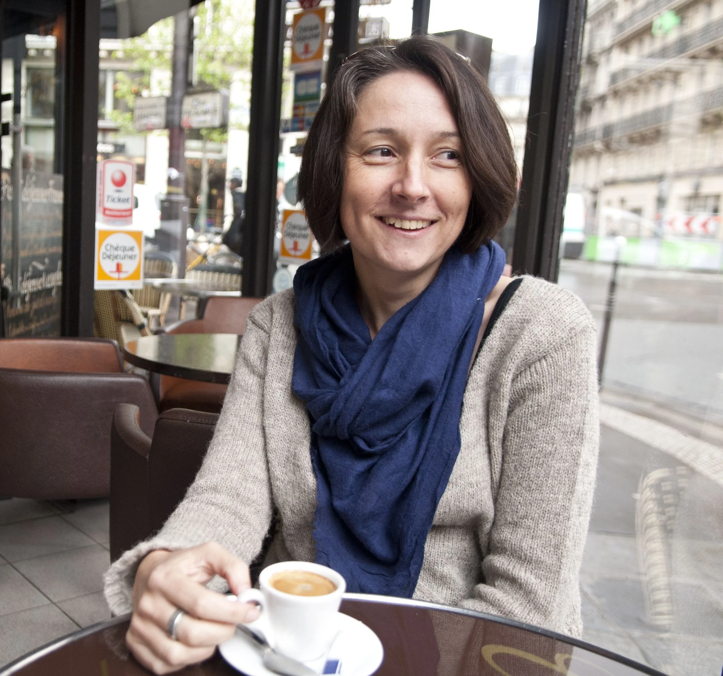 A woman with short brown hair and a beige sweater holding a small white cup of espresso. She is sitting at a dark brown table inside a café, smiling. The background shows outdoor scenery with buildings and street signs.