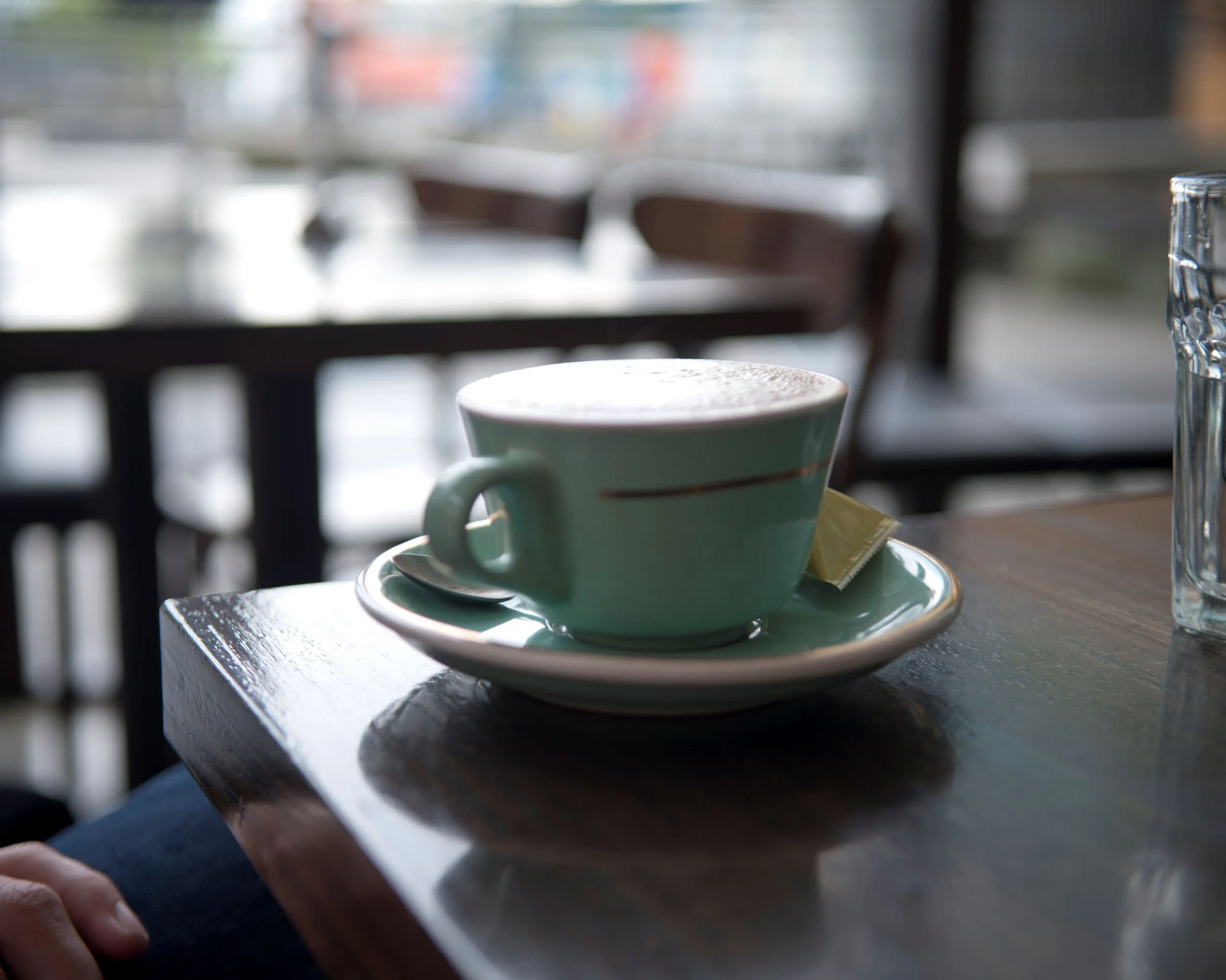 Close-up of a person's hand holding a spoon next to a coffee cup on a saucer, placed on a wooden table in a cafe with blurred background.