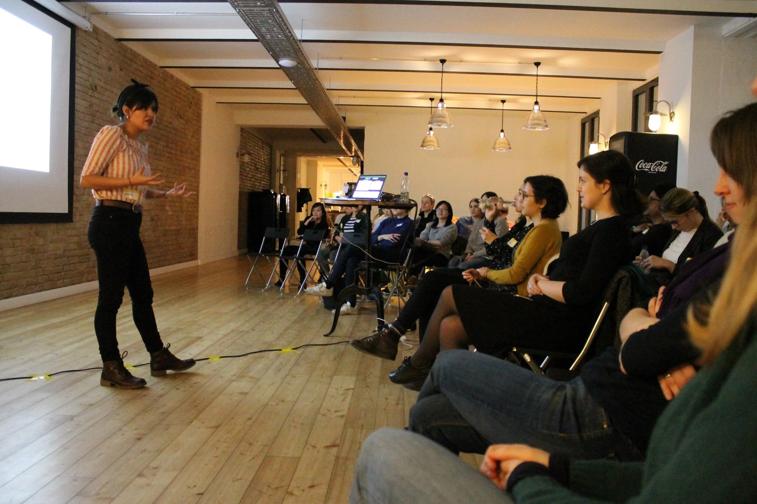 A woman with dark hair in a bun, wearing a striped shirt and black pants, is giving a presentation to an audience seated in a room with brick walls and hanging lights. The audience consists mostly of women, some with glasses, listening attentively.