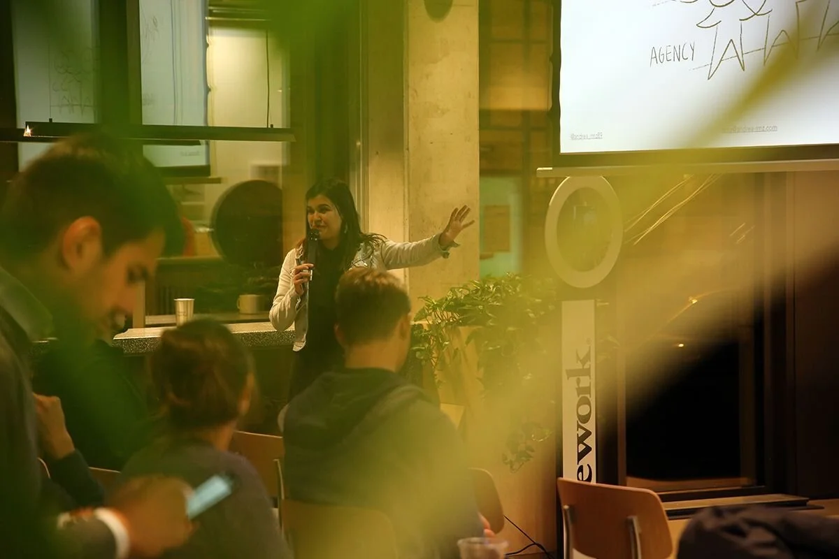 A woman giving a presentation in a room with a group of people seated and listening, some using their phones, and a large screen displaying a slide.