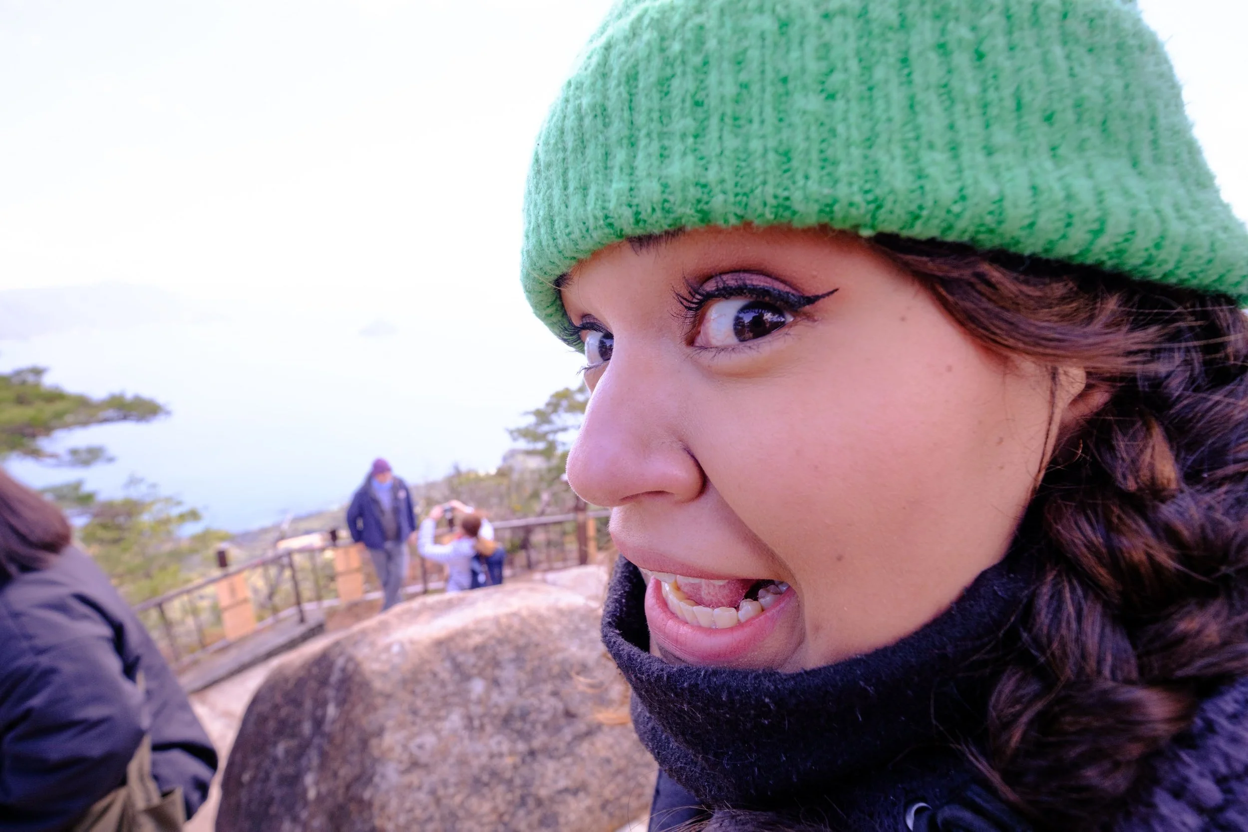 A woman with dark curly hair, makeup, and a green knit hat smiling and looking to the side outdoors with a body of water and people in the background.