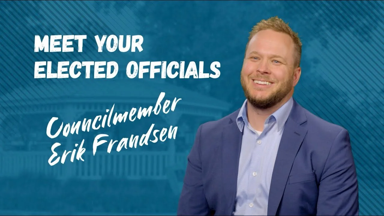 A smiling man in a blue suit with a light blue shirt, standing in front of a background with text that reads, "Meet your elected officials, Councilmember Erik Frandsen."