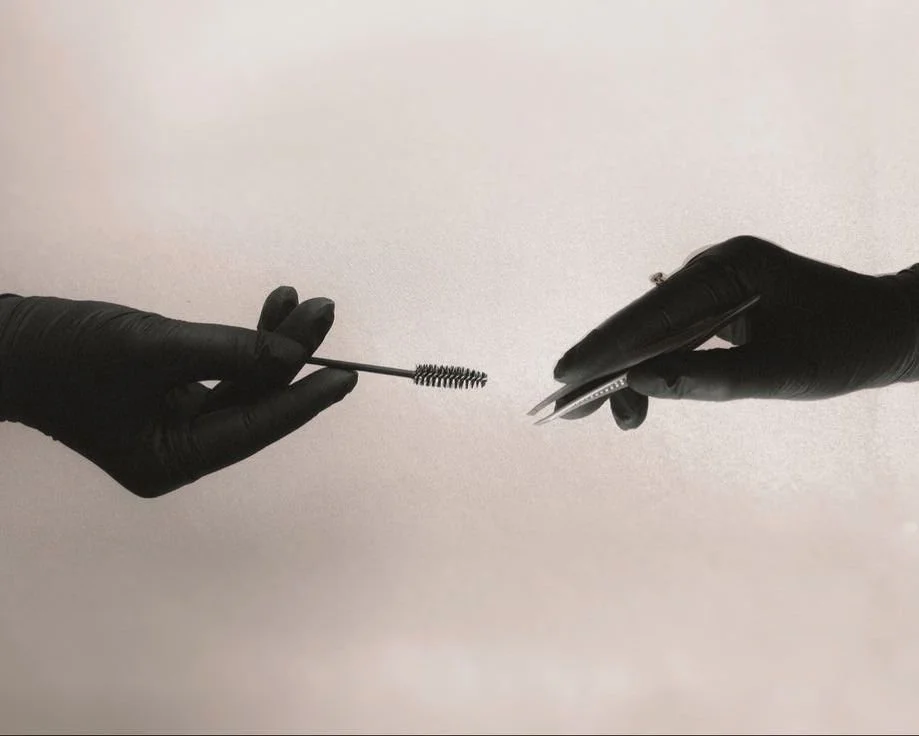 Two hands wearing black gloves, one holding a mascara wand and the other holding a pair of tweezers against a plain background.