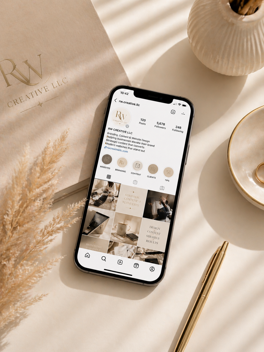 A smartphone displaying the Instagram profile of RW Creative LLC, with branding and website content posts, placed on a beige surface alongside a gold pen, a ceramic vase, and a bag with the RW Creative logo.