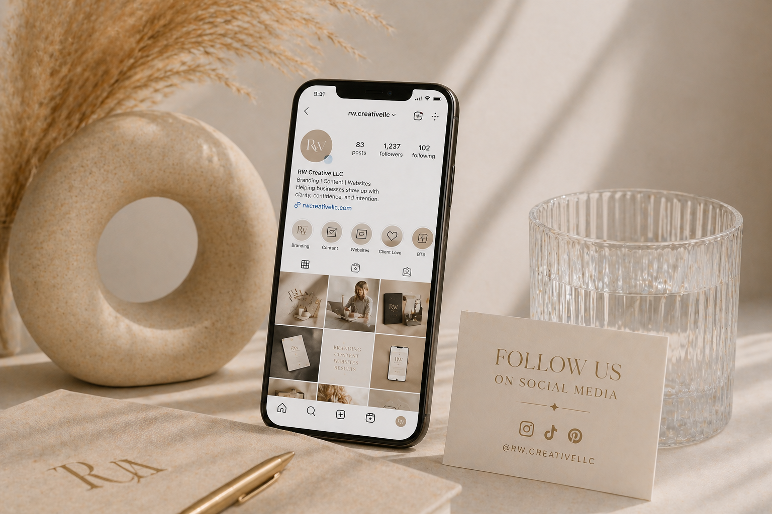 Instagram profile page on an iPhone, placed on a beige surface. To the right, a textured glass of water and a social media promotion card with @RW.CREATIVELLc. Behind the phone, a beige decorative ring and a beige notebook with 'RWA' logo, along with a gold pen.