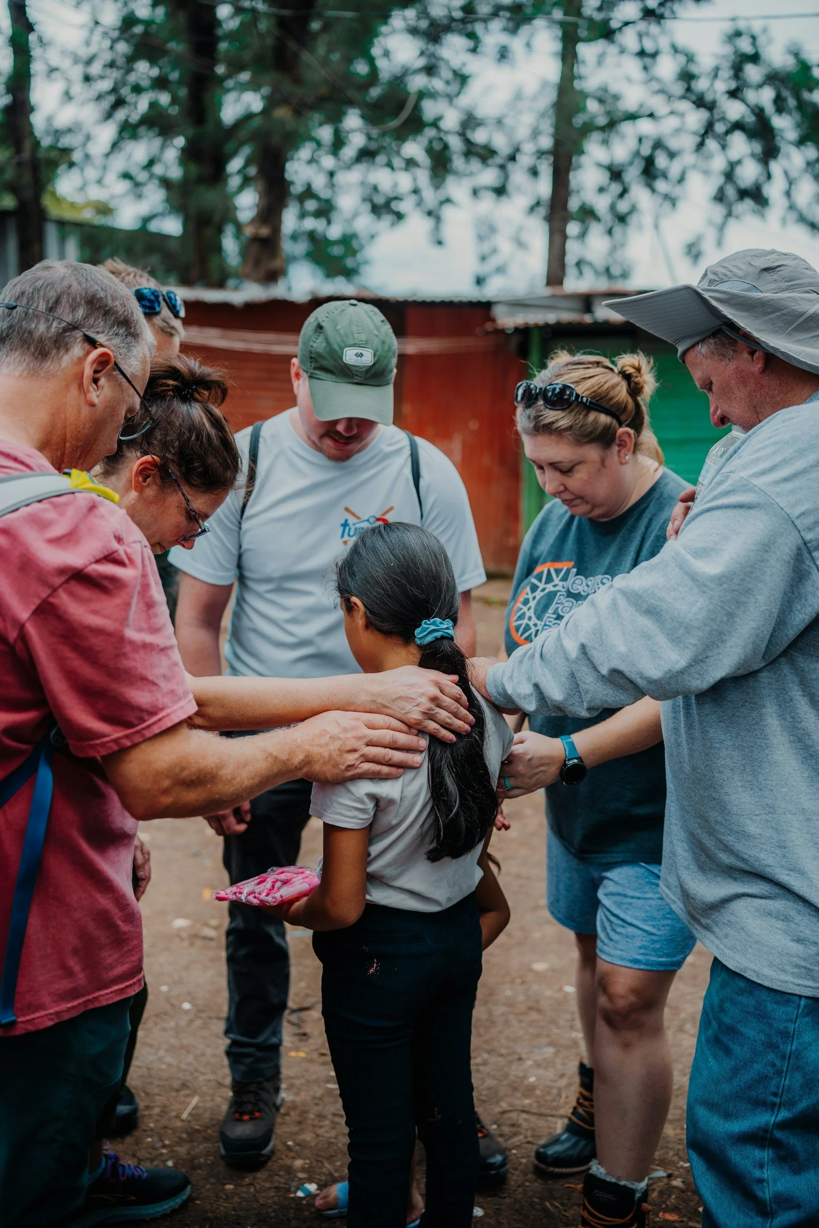 Group of people surrounding a young girl in an outdoor setting, appearing to pray or have a moment of reflection.