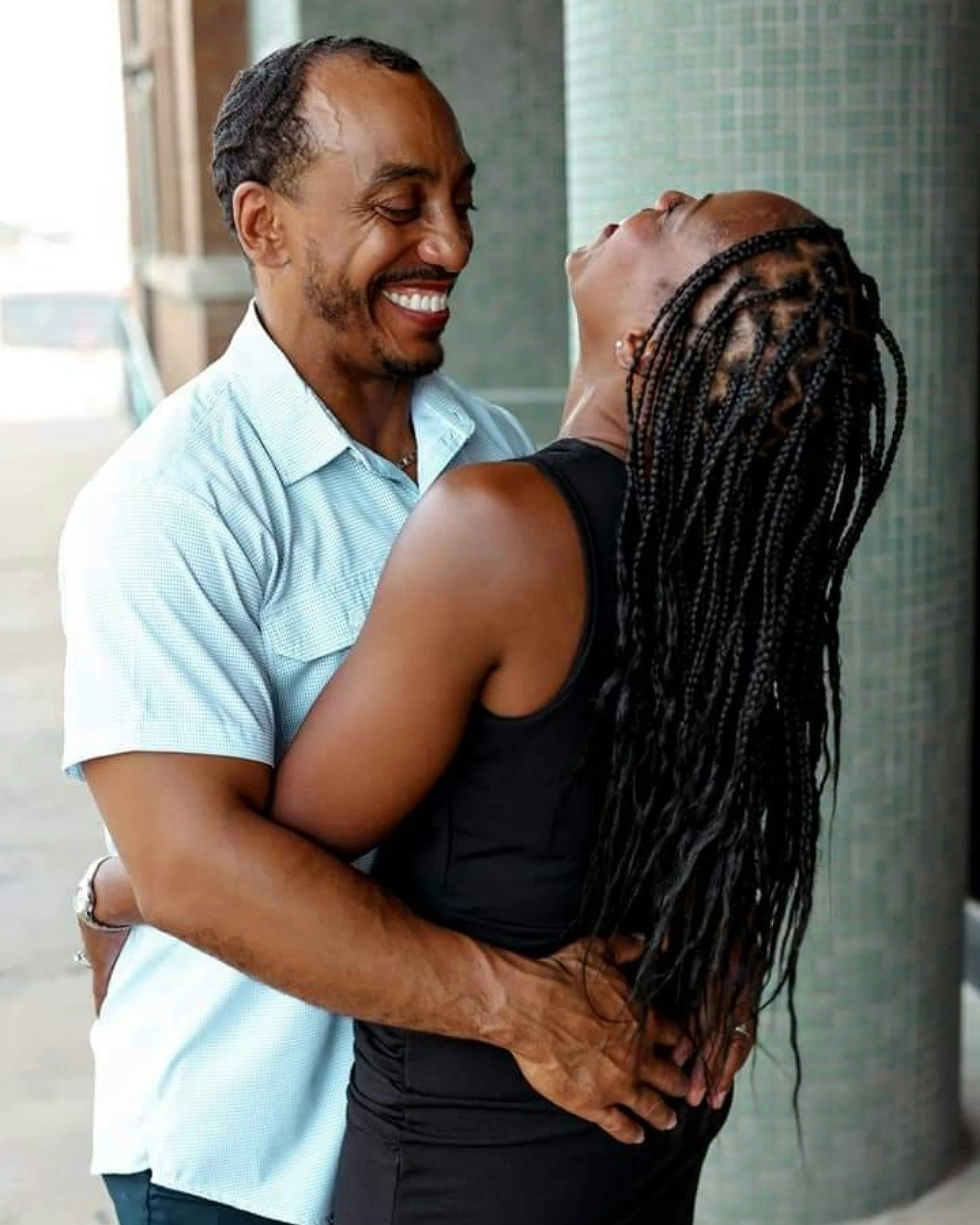 A man and a woman sharing a joyful embrace, with the man smiling visible. The man has a beard and is wearing a light blue shirt, and the woman has braided hair and is wearing a black sleeveless top.