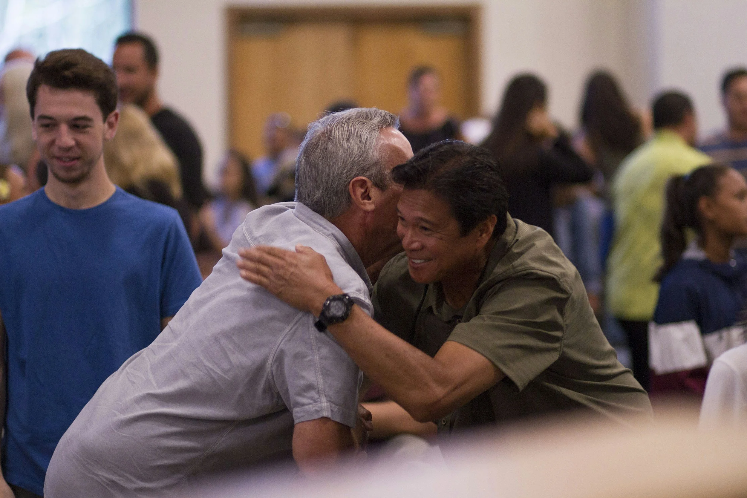 Two men are hugging and greeting each other warmly in a crowded indoor setting, possibly a community center or church.