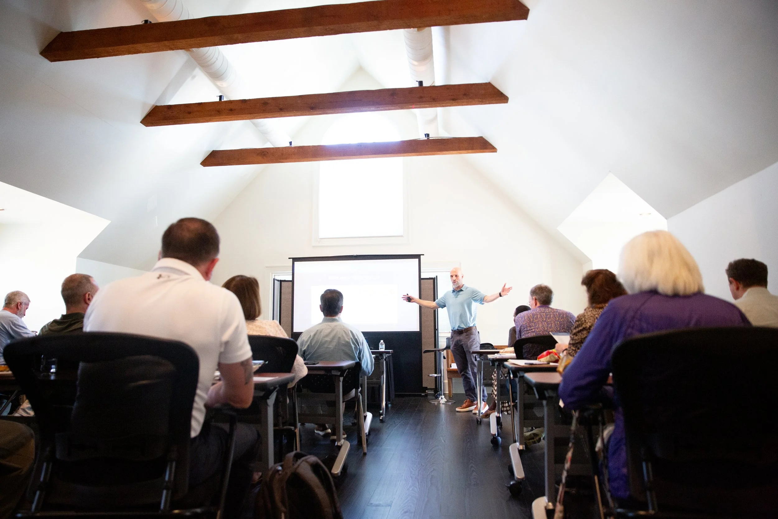 A presenter stands in front of a room full of seated audience members during a conference or seminar in a bright, spacious room with a large window and exposed wooden beams on the ceiling.