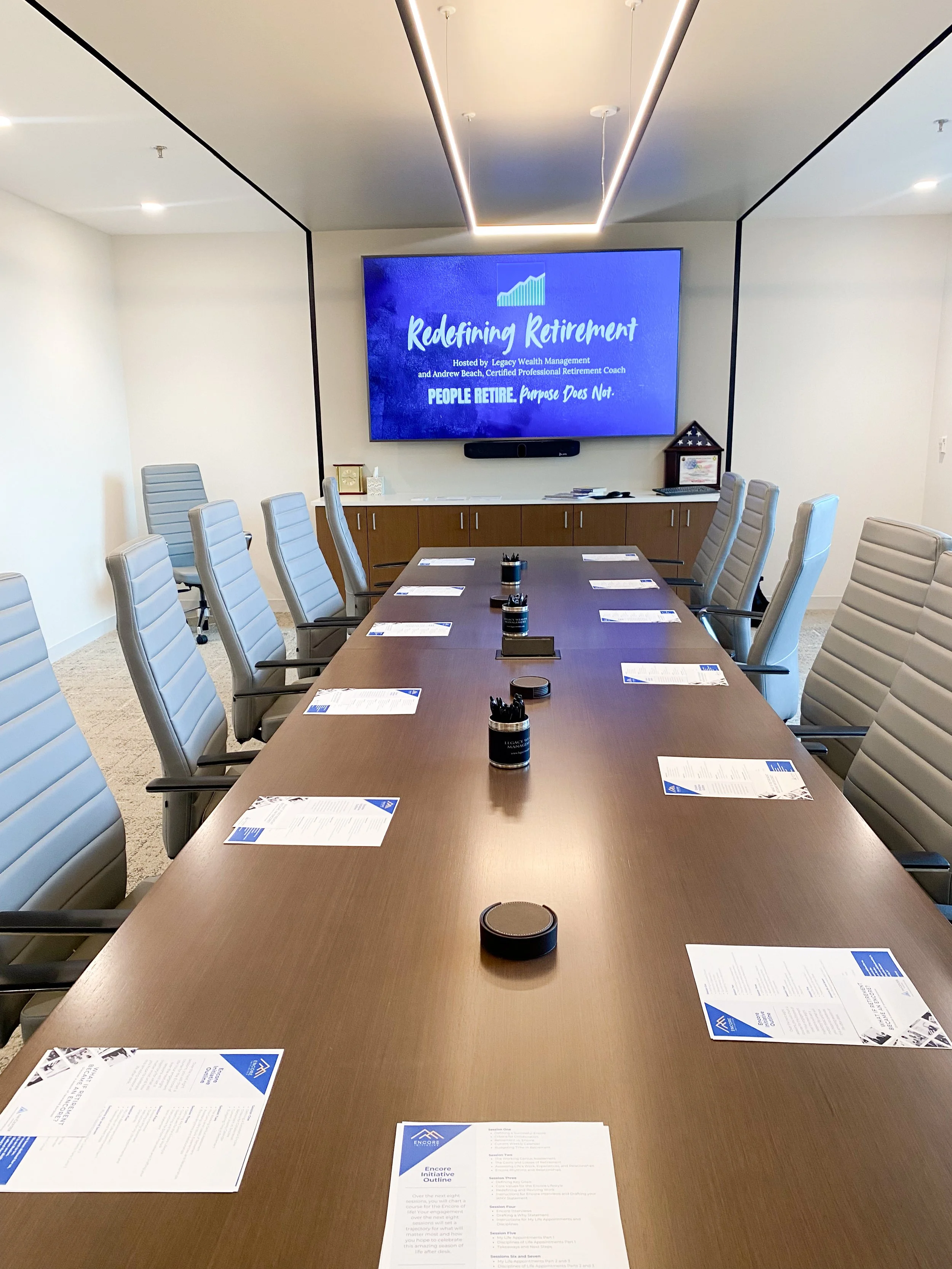 A conference room with a large wooden table surrounded by gray office chairs. The table has papers, pens, and round black containers on it. A large screen at the front displays a presentation titled 'Redefining Retirement.' The room has white walls, a ceiling with recessed lighting, and a cabinet with decorative items at the back.
