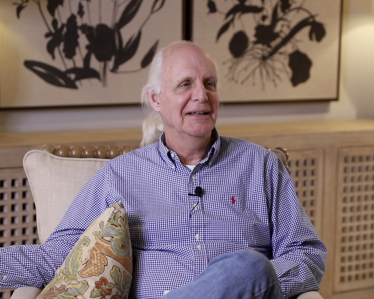 An older man with long white hair sitting on a beige armchair with a colorful pillow, in a room with artwork featuring black abstract shapes on the wall behind him.