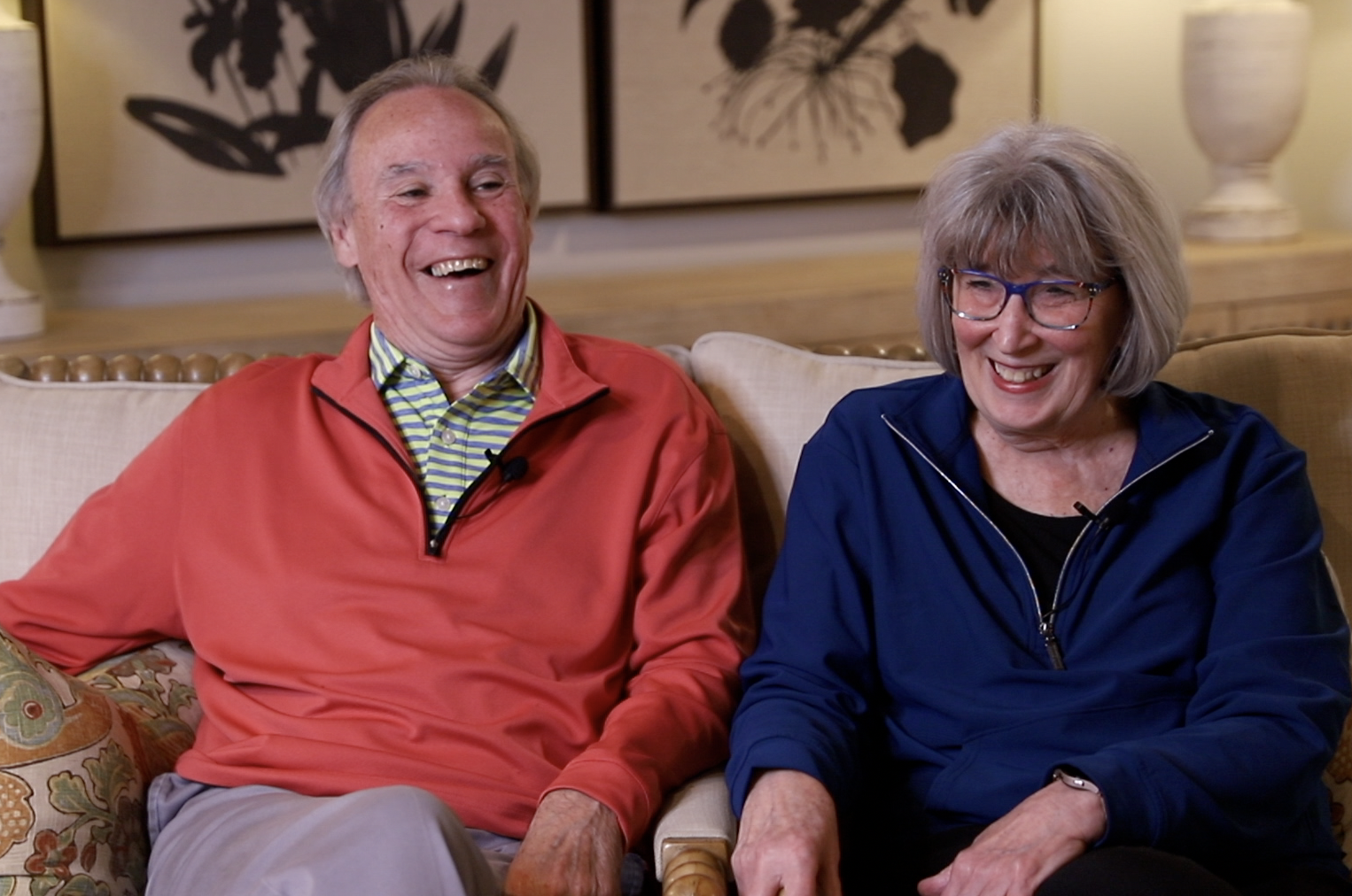 Two elderly people, a man in a red jacket and a woman in a blue jacket, sitting on a beige sofa in a warmly decorated living room, laughing and enjoying each other's company.
