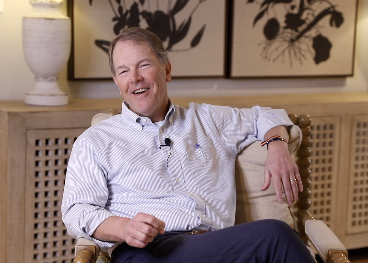 A middle-aged man with light brown hair sitting on a wooden chair, smiling and wearing a white striped button-up shirt with the sleeves rolled up, in a room with beige walls, a wooden cabinet, and black and white floral artwork on the wall behind him.