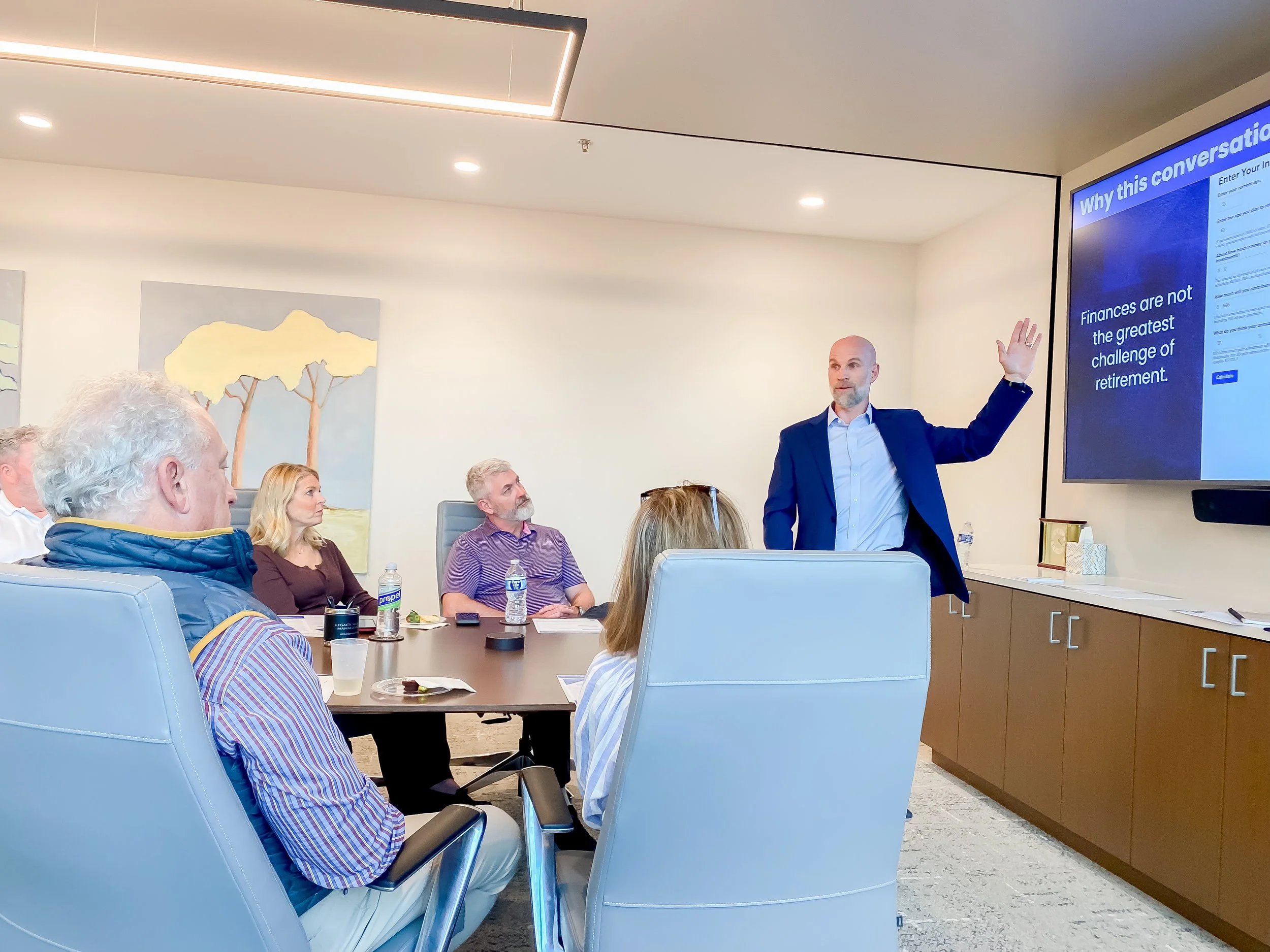 A man in a blue suit giving a presentation to a group of people in a conference room, with a large screen displaying information about financial challenges in retirement.