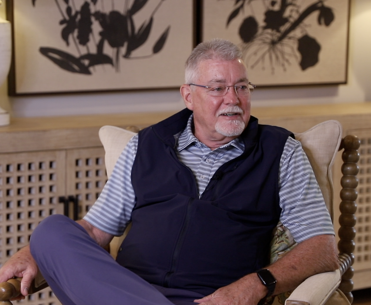 An elderly man with glasses and a white beard sitting on a beige armchair, wearing a striped shirt and a navy vest, in a room with artwork of black floral silhouettes on the wall behind him.