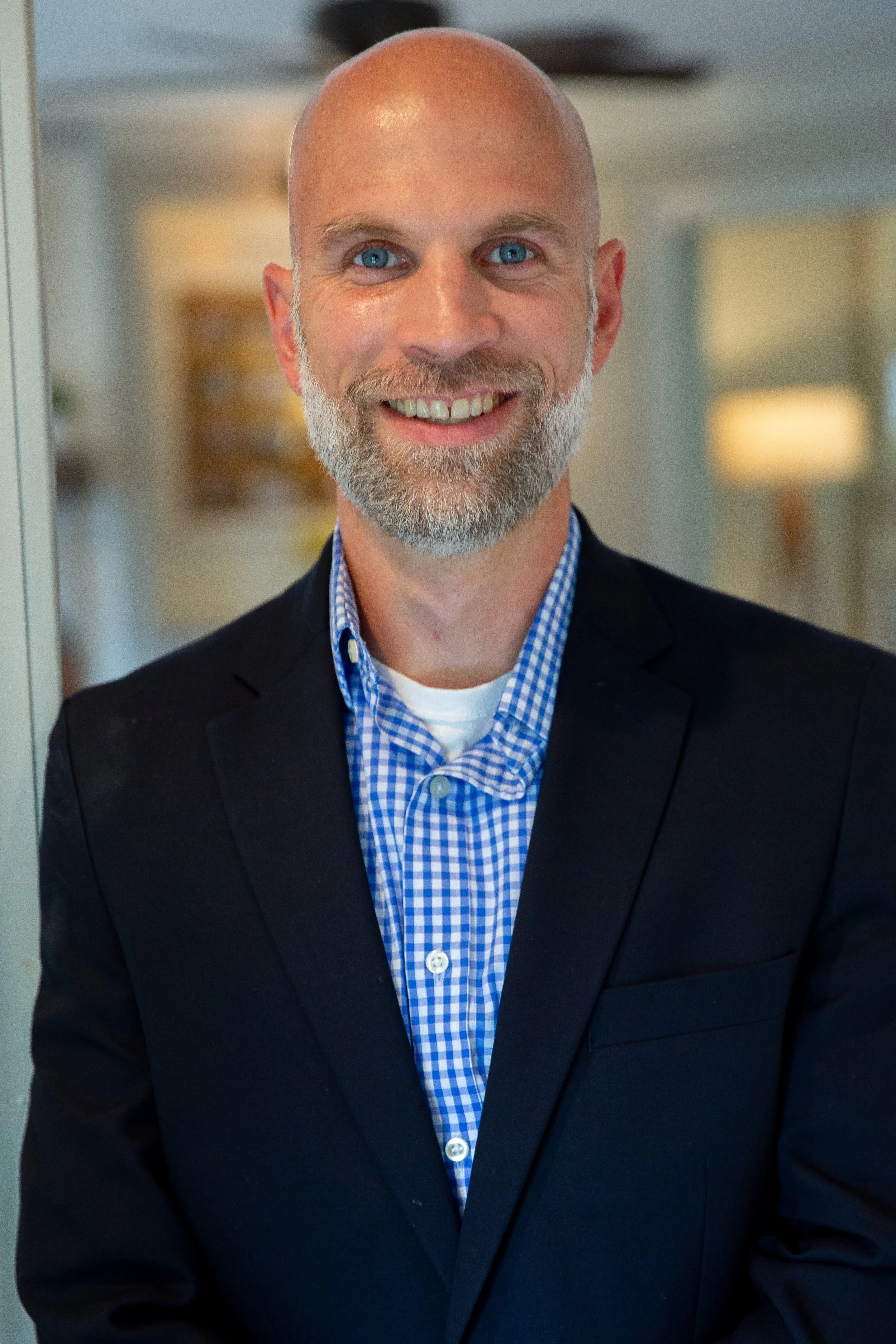 Smiling middle-aged man with a bald head, blue eyes, and a beard, wearing a dark suit jacket over a blue checkered shirt, standing in an indoor setting.
