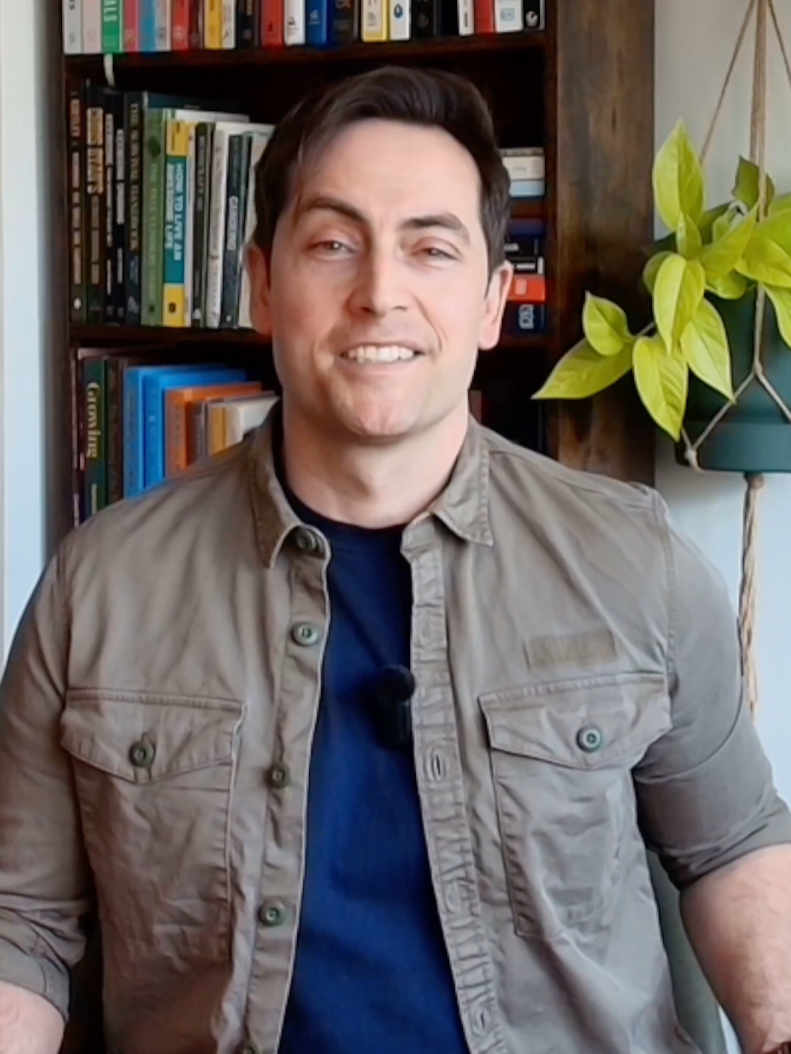 A man with dark hair smiling while sitting in front of a bookcase filled with books, next to a potted plant with green leaves.