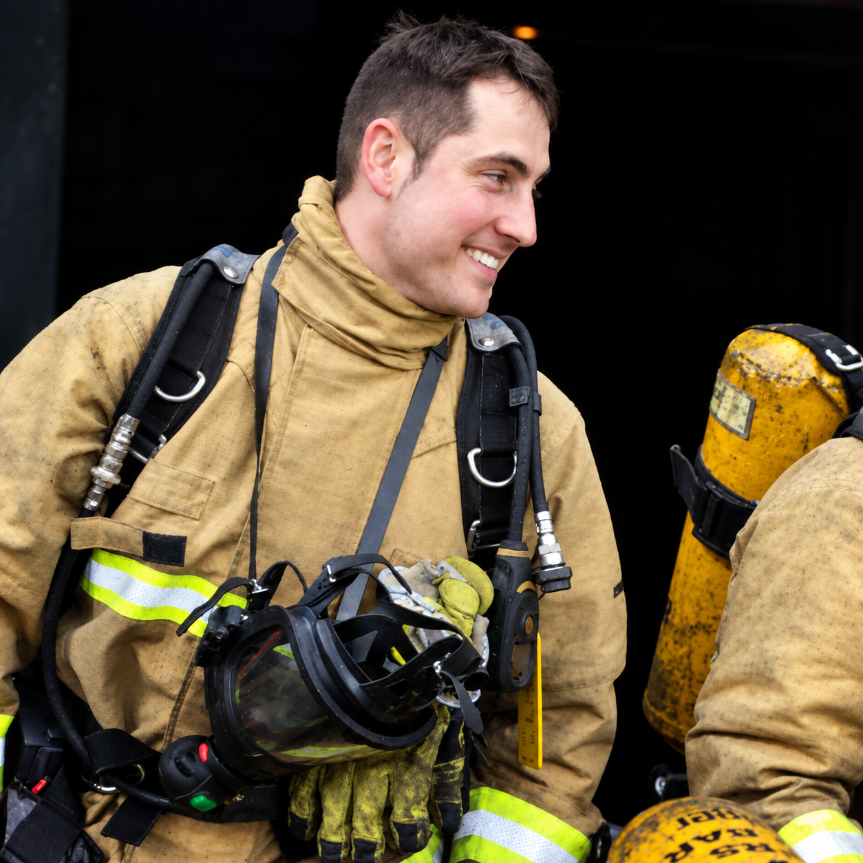 A smiling firefighter wearing tan turnout gear and a face mask hanging from his neck, with protective gloves and a yellow air tank on his back.