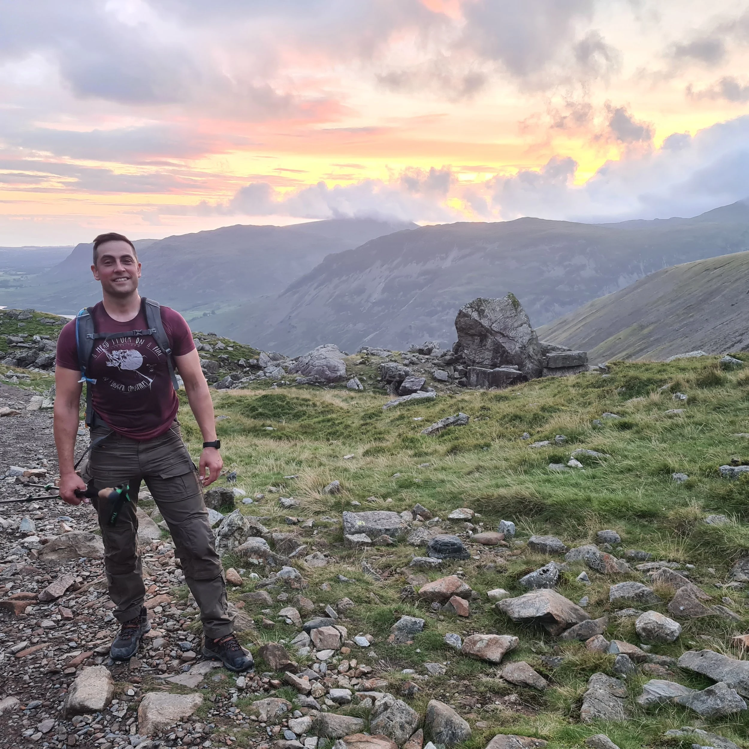 A young man standing outdoors on rocky terrain with mountains and a colorful sunset sky in the background.