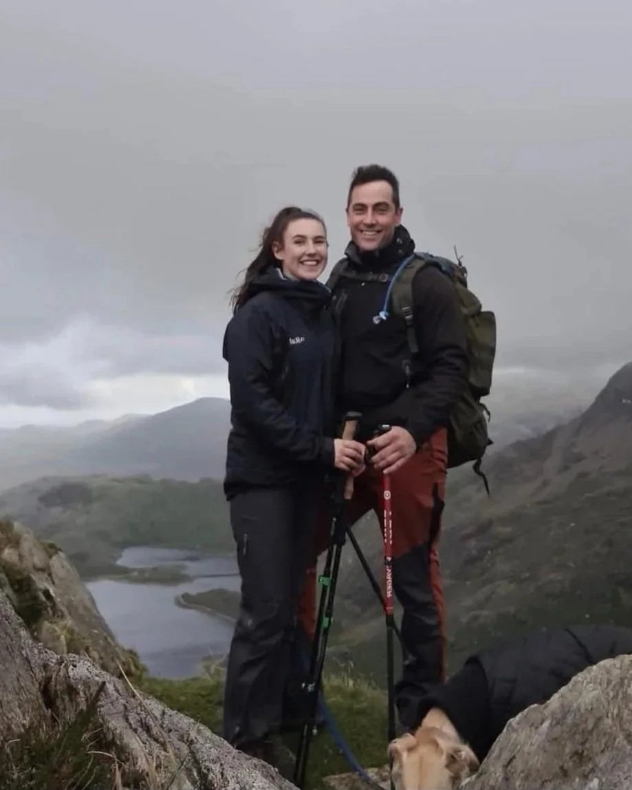 A smiling couple hiking outdoors in a mountainous area with a lake in the background, holding hiking poles, along with a small dog on a leash.